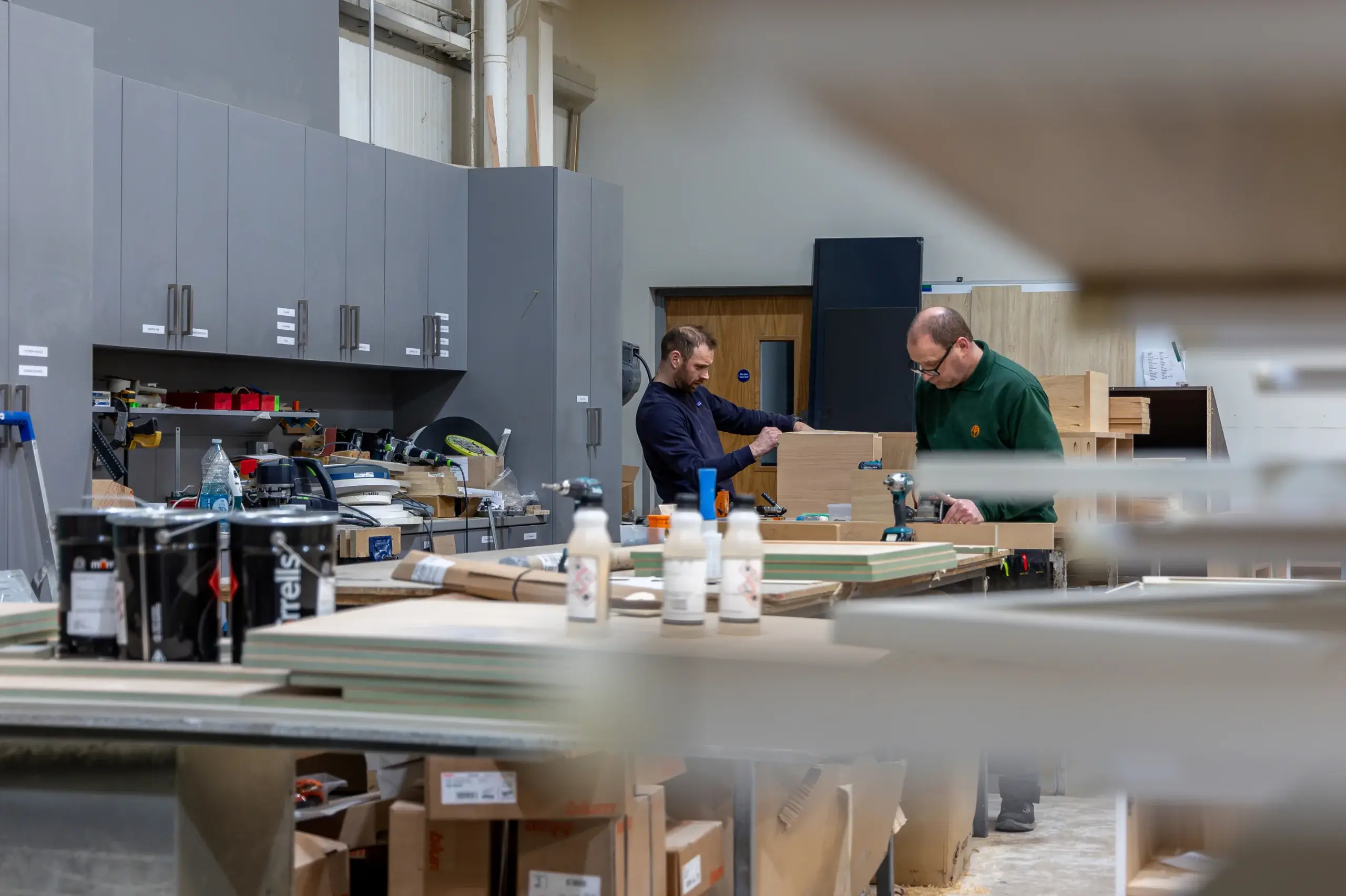 Two men assembling wooden cabinets in a cluttered workshop with tools, paint cans, and stacked panels on workbenches.
