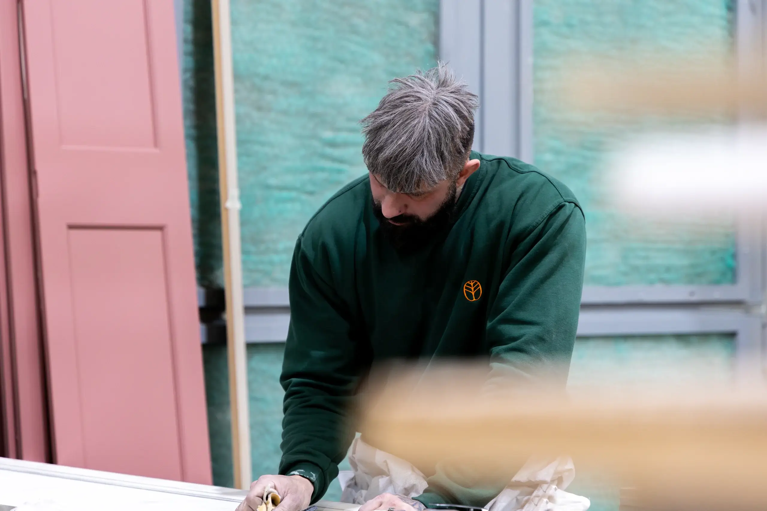 Man with gray-streaked hair and beard leaning over a workbench, wearing a dark green sweatshirt with a small orange leaf logo, sanding or working on wood.