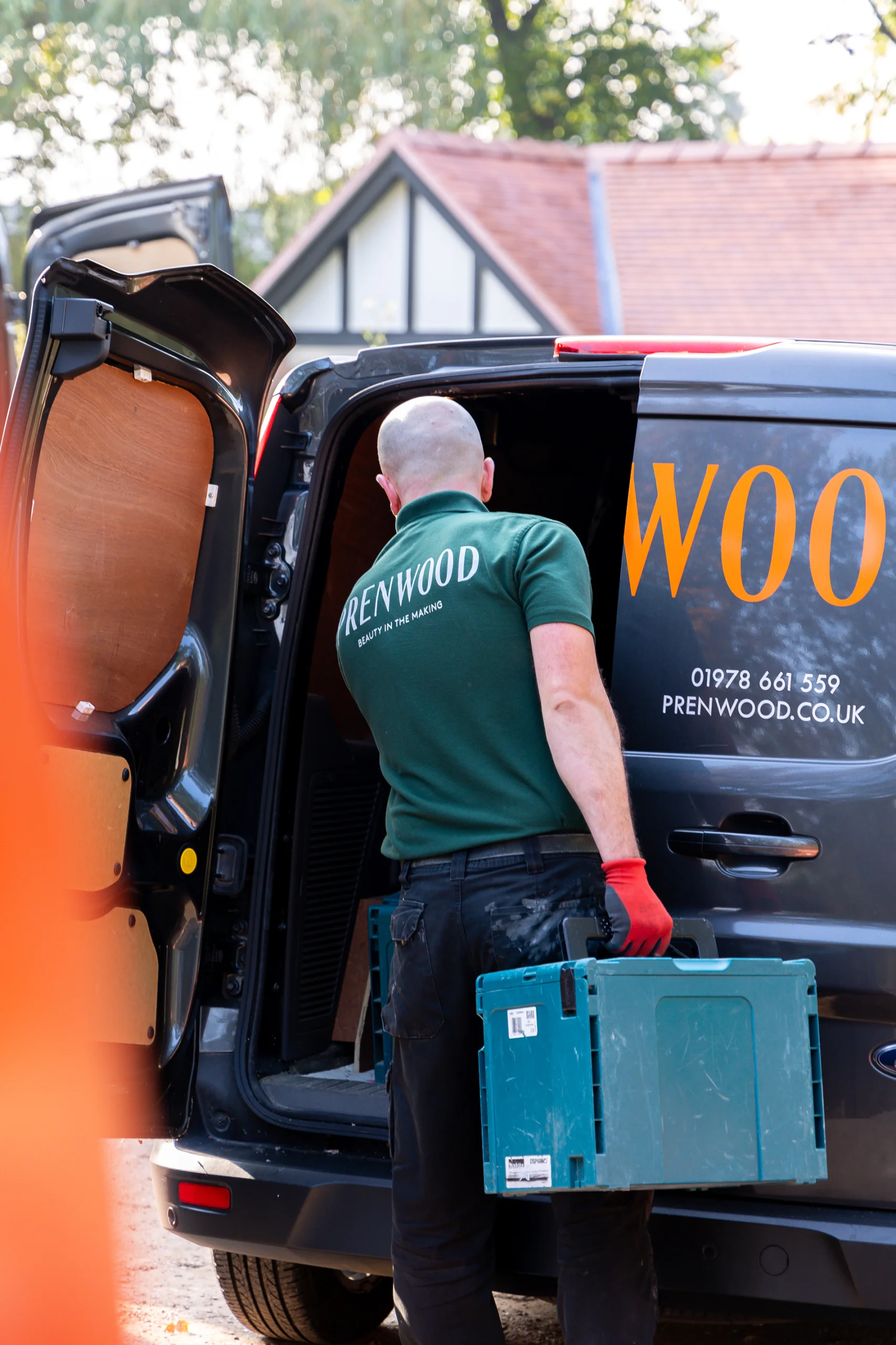 Worker in green "Prenwood" polo carrying a teal toolbox into a black company van with orange lettering.