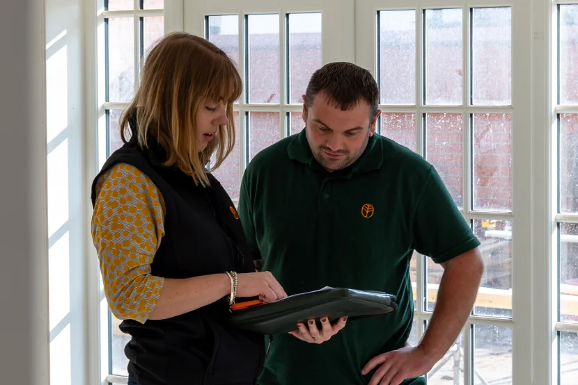 A man and woman stand by a window, both focused on a tablet in their hands, engaged in conversation.