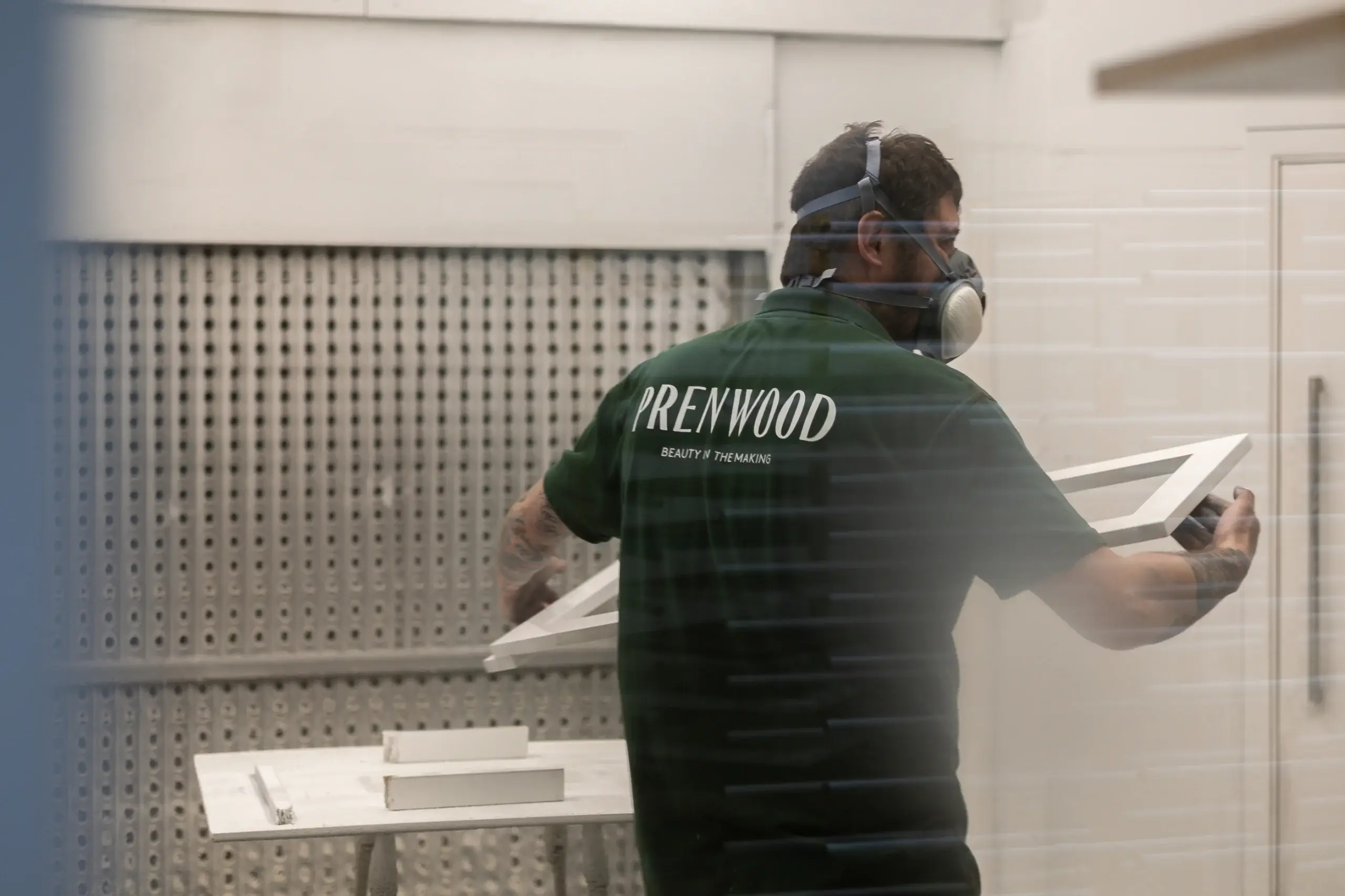Worker wearing respirator and green "Prenwood" shirt holding a painted white frame inside a workshop