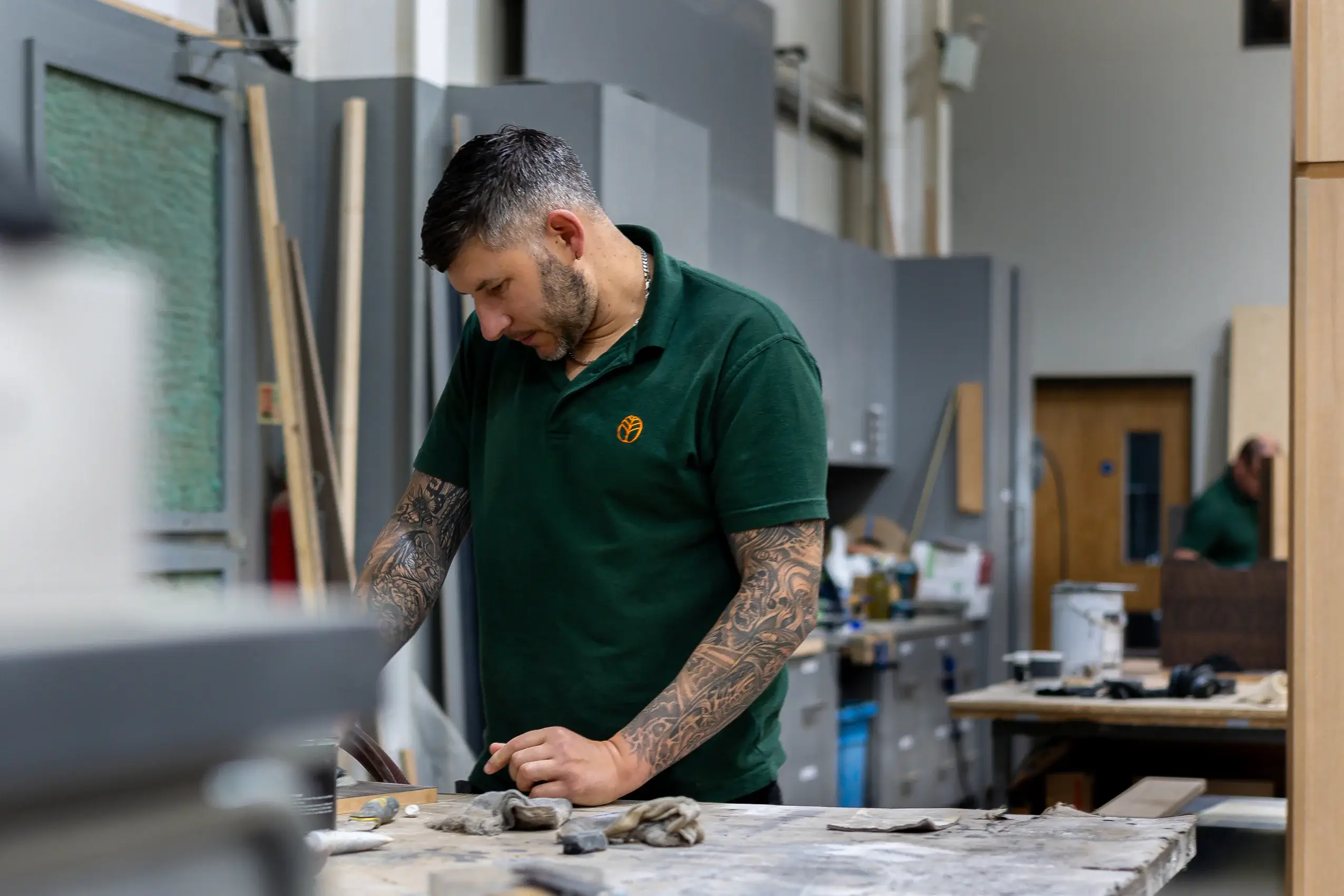 Man with full-sleeve tattoos leaning over a workshop table, inspecting or working on a small object in a green polo shirt.