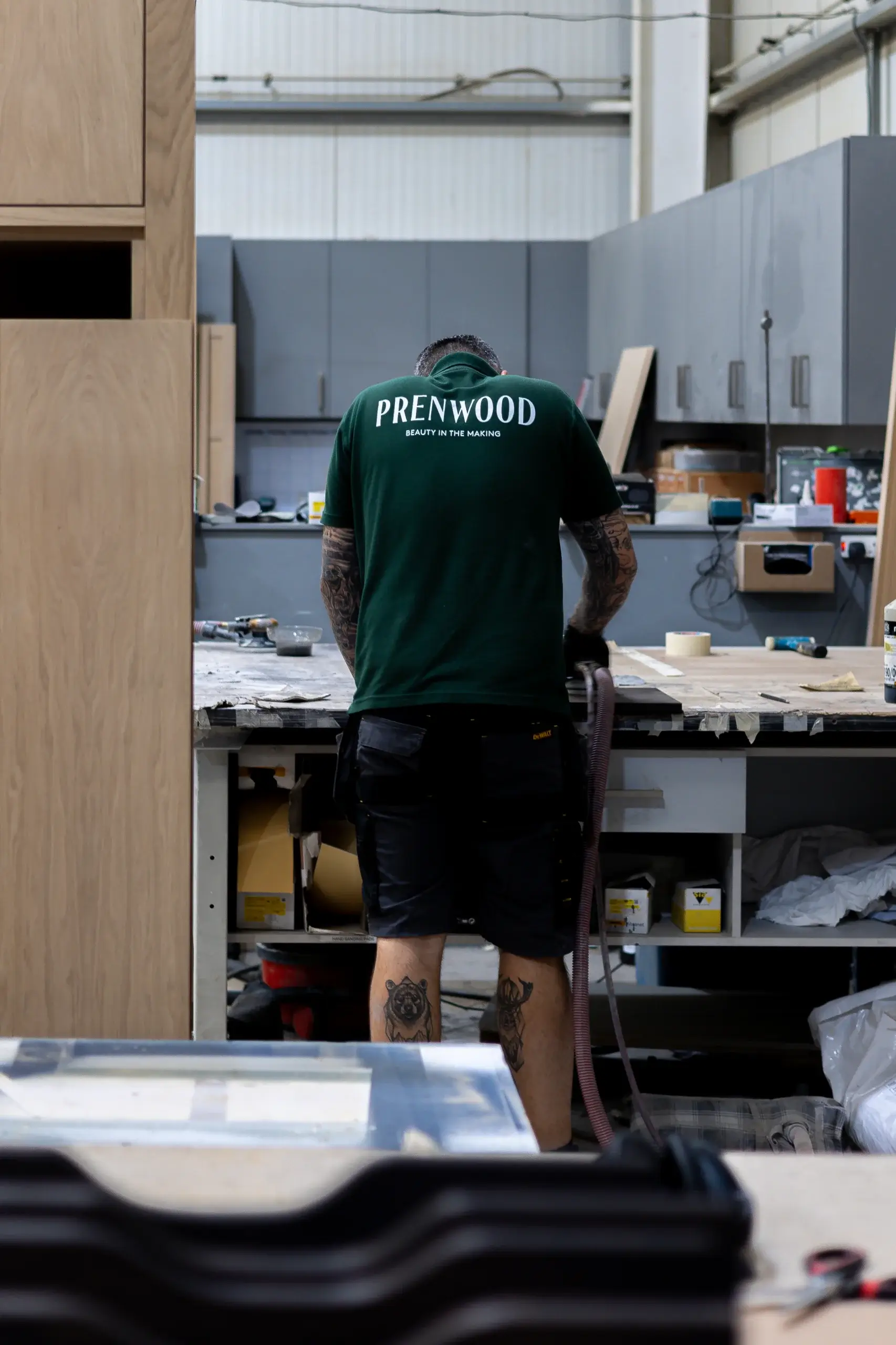 Man in green "PRENWOOD" shirt working at a cluttered woodshop bench, visible tattoos on forearms and calves.