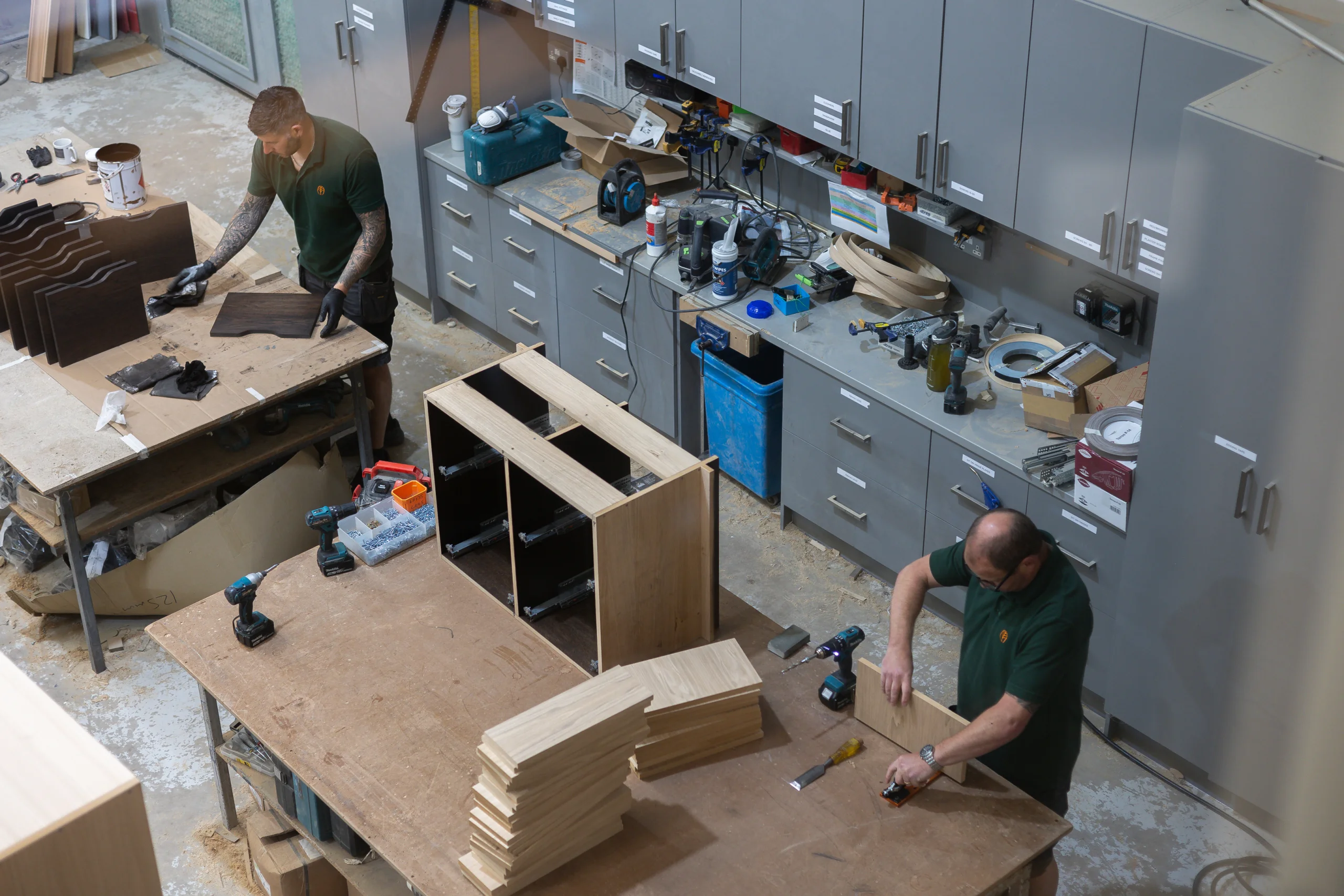 Two men in green shirts assembling wooden cabinet parts on large workbenches in a busy carpentry workshop