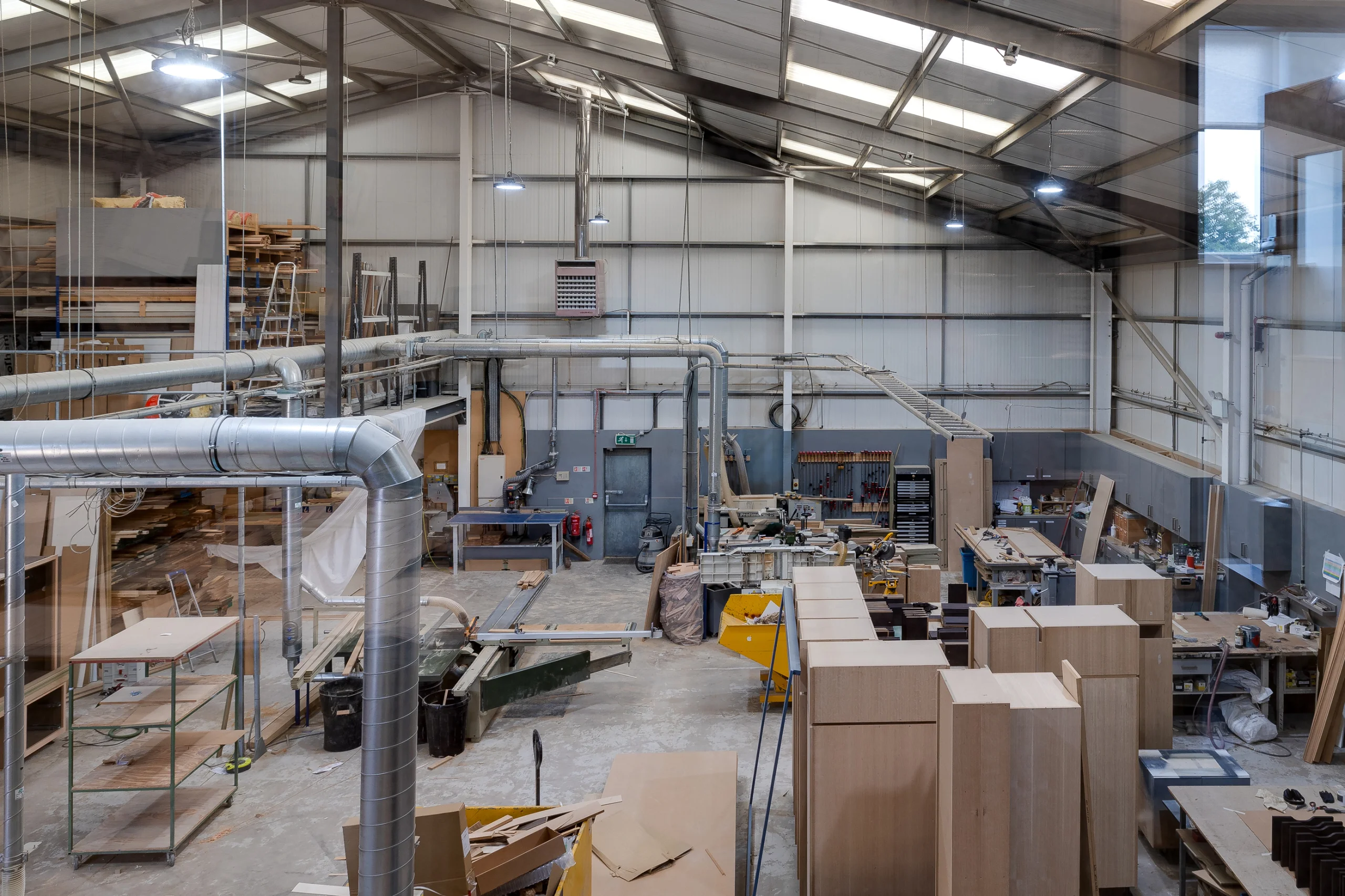 Spacious woodworking shop with tall ventilation ducts, unfinished wooden cabinets, workbenches, tools, and overhead lighting