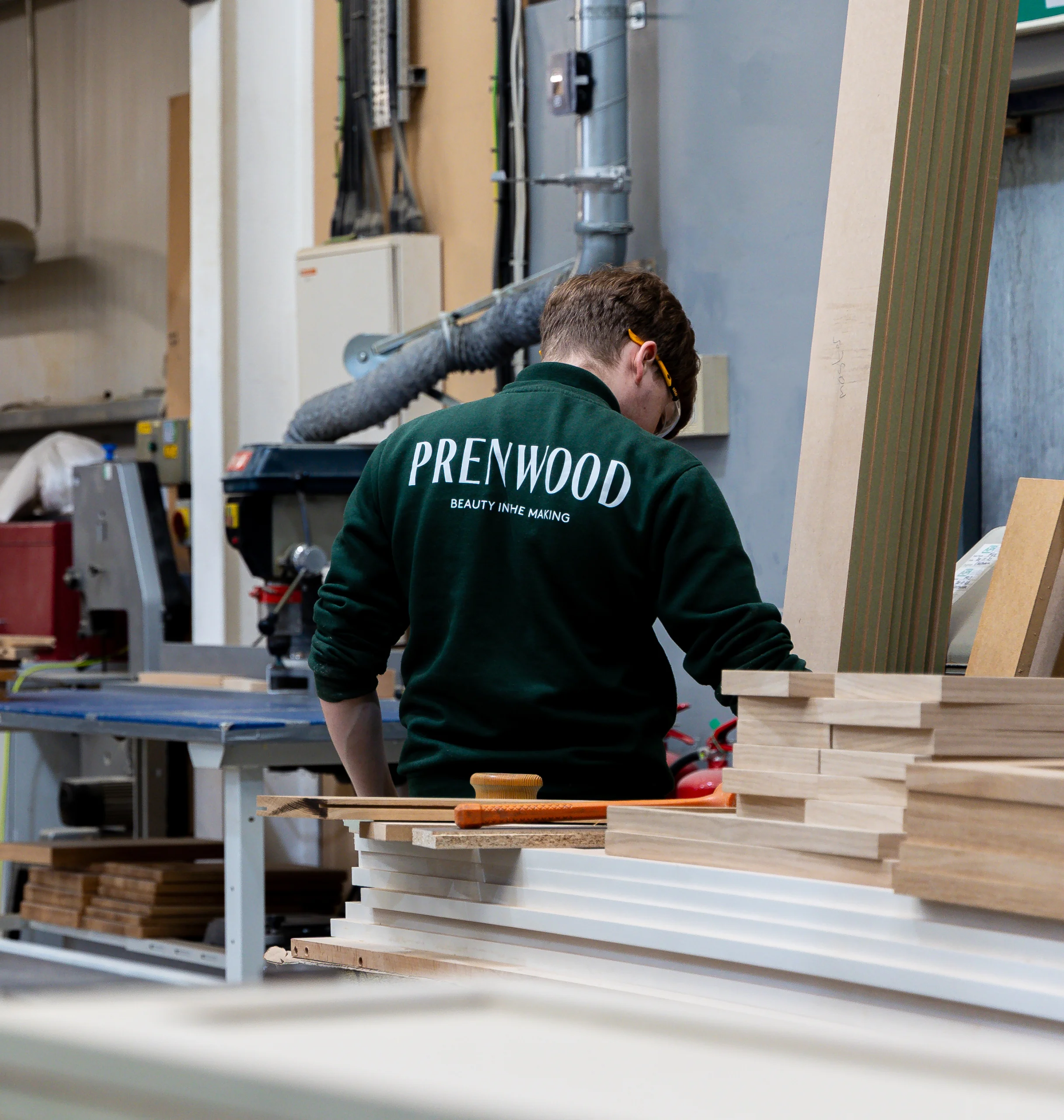 Person in green "PRENWOOD" sweatshirt working in a woodworking shop, standing near stacked wooden boards and tools.