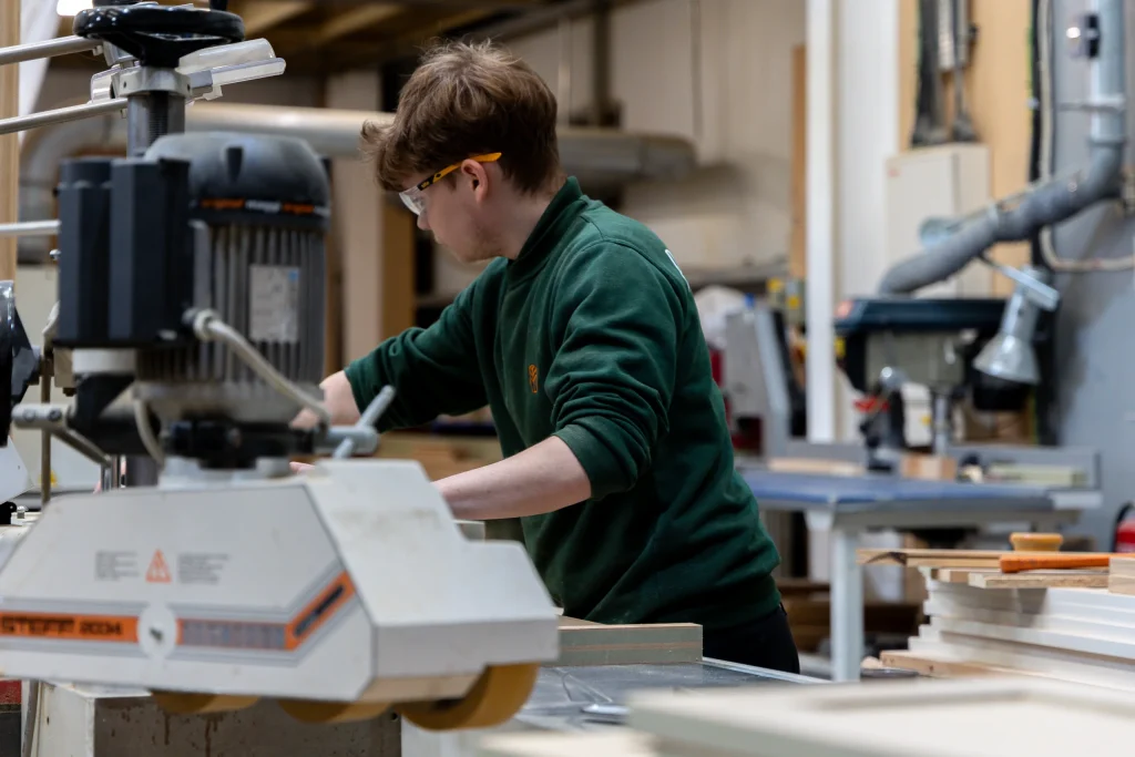 Young person wearing safety glasses and a green sweatshirt operating a woodworking machine in a workshop, surrounded by lumber and tools.
