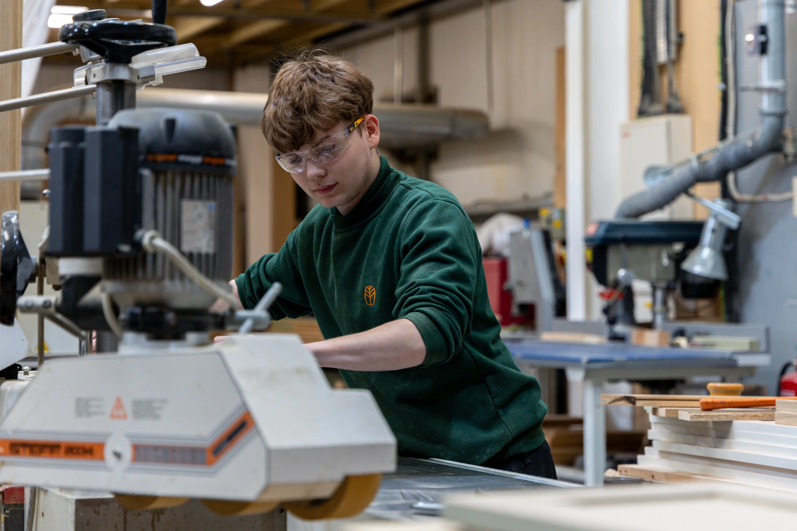 Young person wearing safety goggles operating a woodworking machine in a busy workshop, concentrating on their task