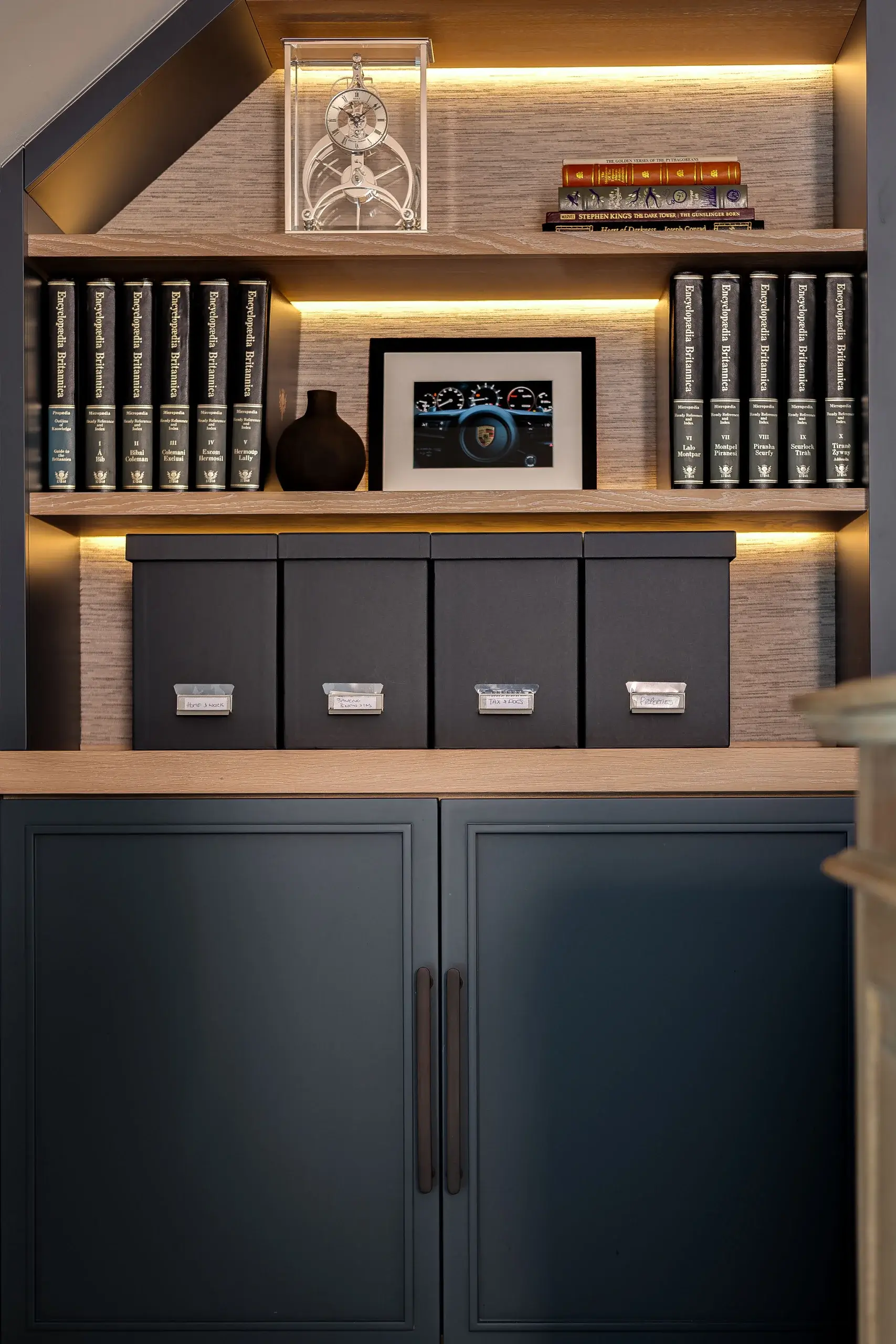 Built-in wooden shelving with backlit shelves holding encyclopedias, decorative clock, framed car dashboard photo, and four black storage boxes.