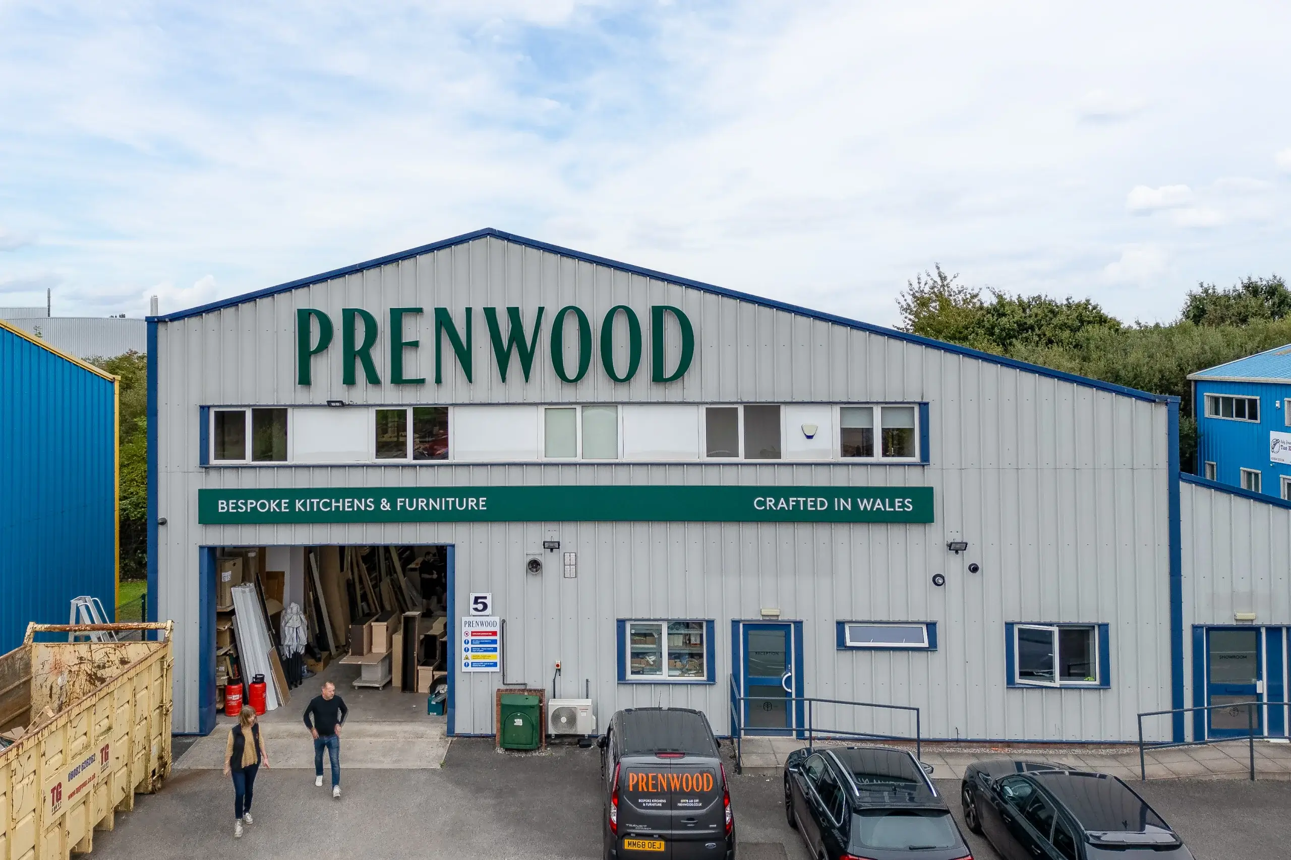 Large light-gray industrial building with green "PRENWOOD" sign, workshop entrance, parked vans and two people walking outside.