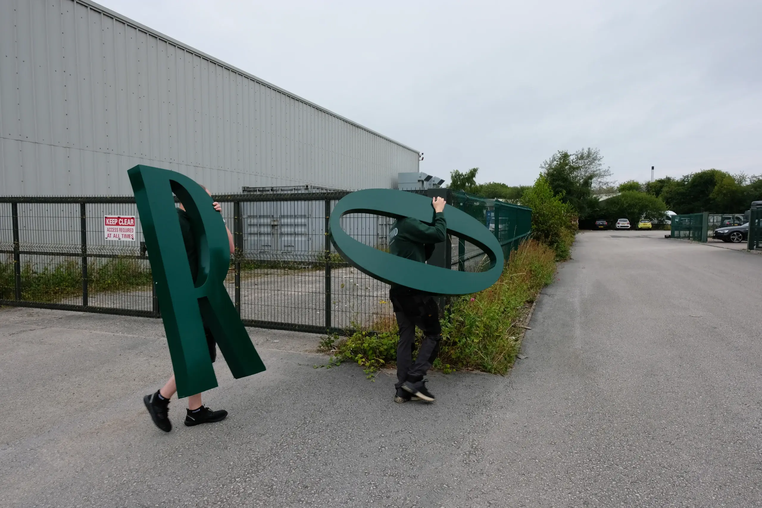 Two people carry oversized dark green sculpted letters (an R and an O) past a fenced industrial yard.