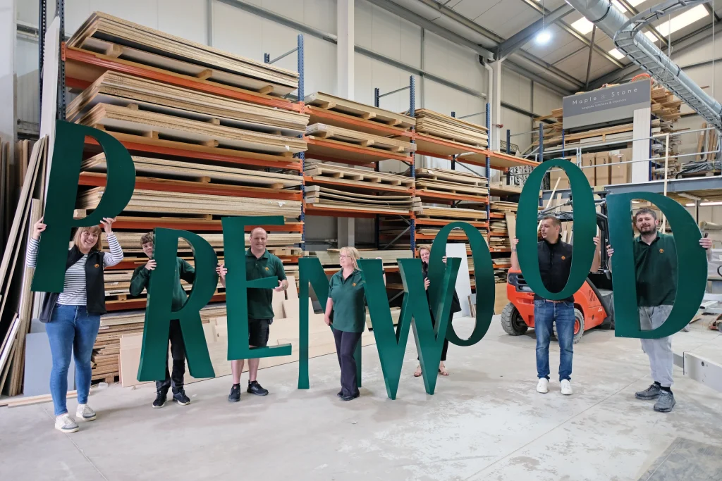 Seven people in a workshop holding large green letters that spell PRENWOOD against stacked wood racks.
