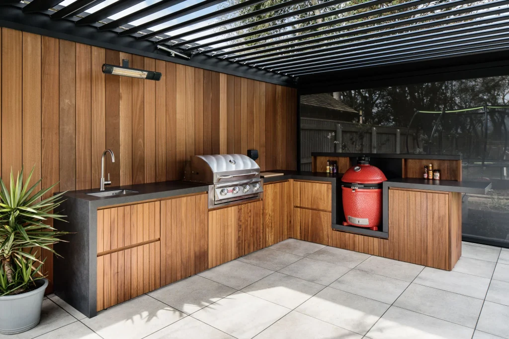 Outdoor modern wood-paneled kitchen with stainless grill, red ceramic kamado smoker, sink, and pergola roof