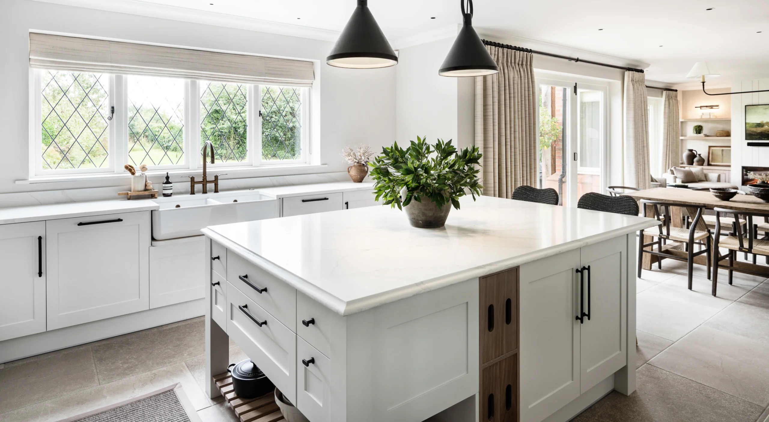 Bright modern kitchen with a large white island topped by a potted green plant, farmhouse sink under diamond-pane windows, and dining area in the background.