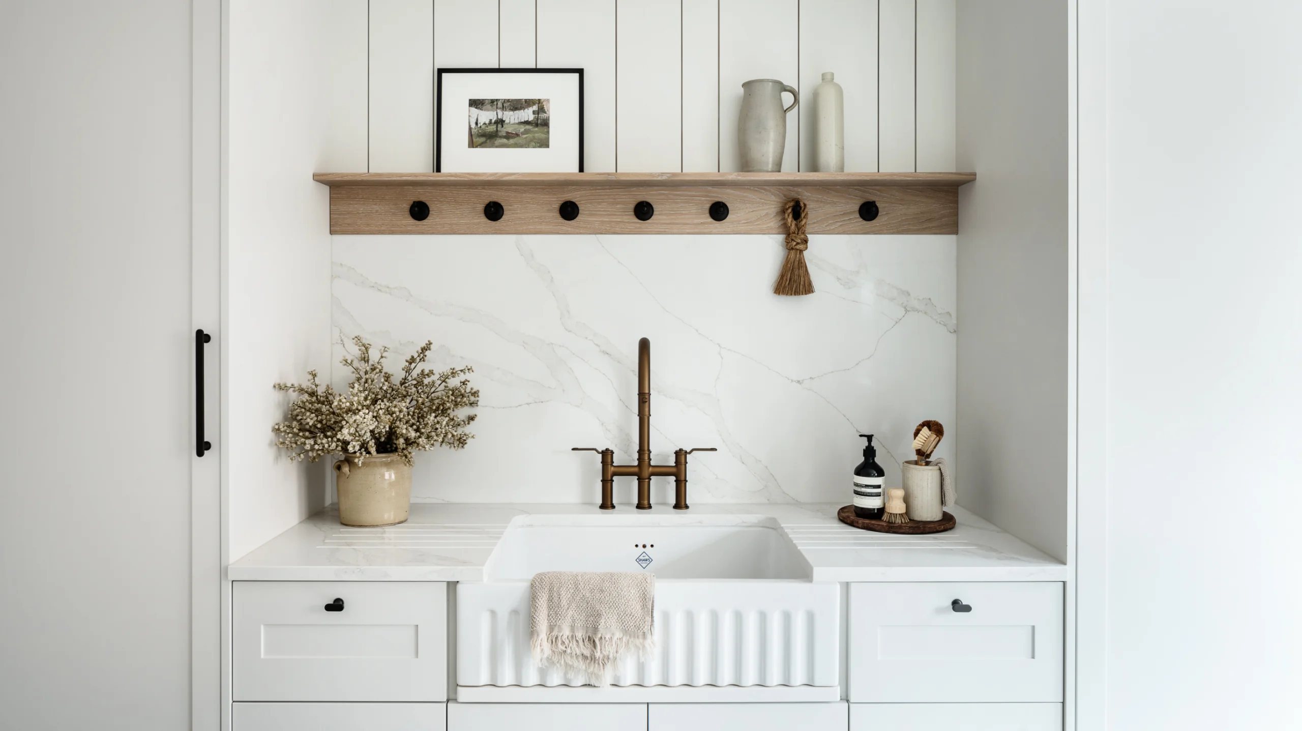 Kitchen sink alcove with a white apron-front sink, brass bridge faucet, marble backsplash, wooden shelf with hooks, vase of dried flowers, and soap accessories.