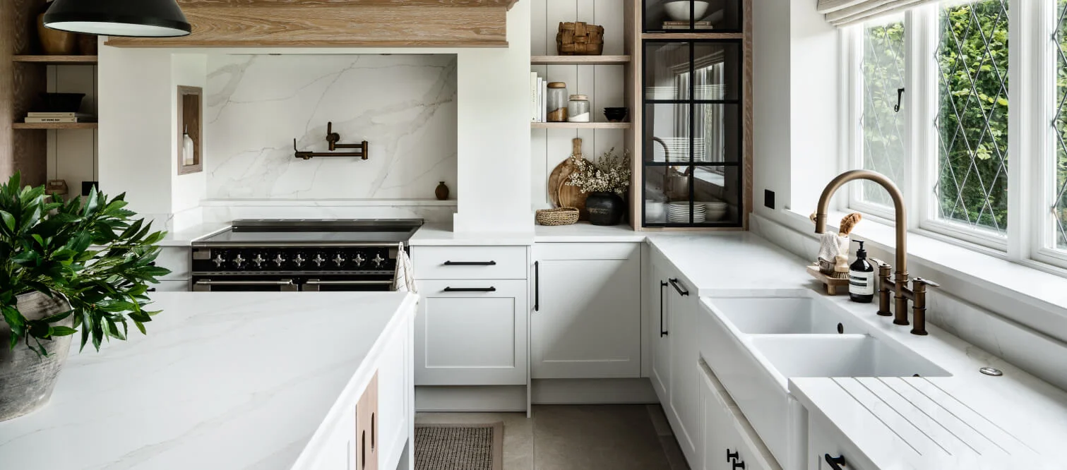 A modern kitchen with white cabinets, a marble backsplash, an oven, a farmhouse sink, and a window. Decor includes plants and pottery on open shelves.