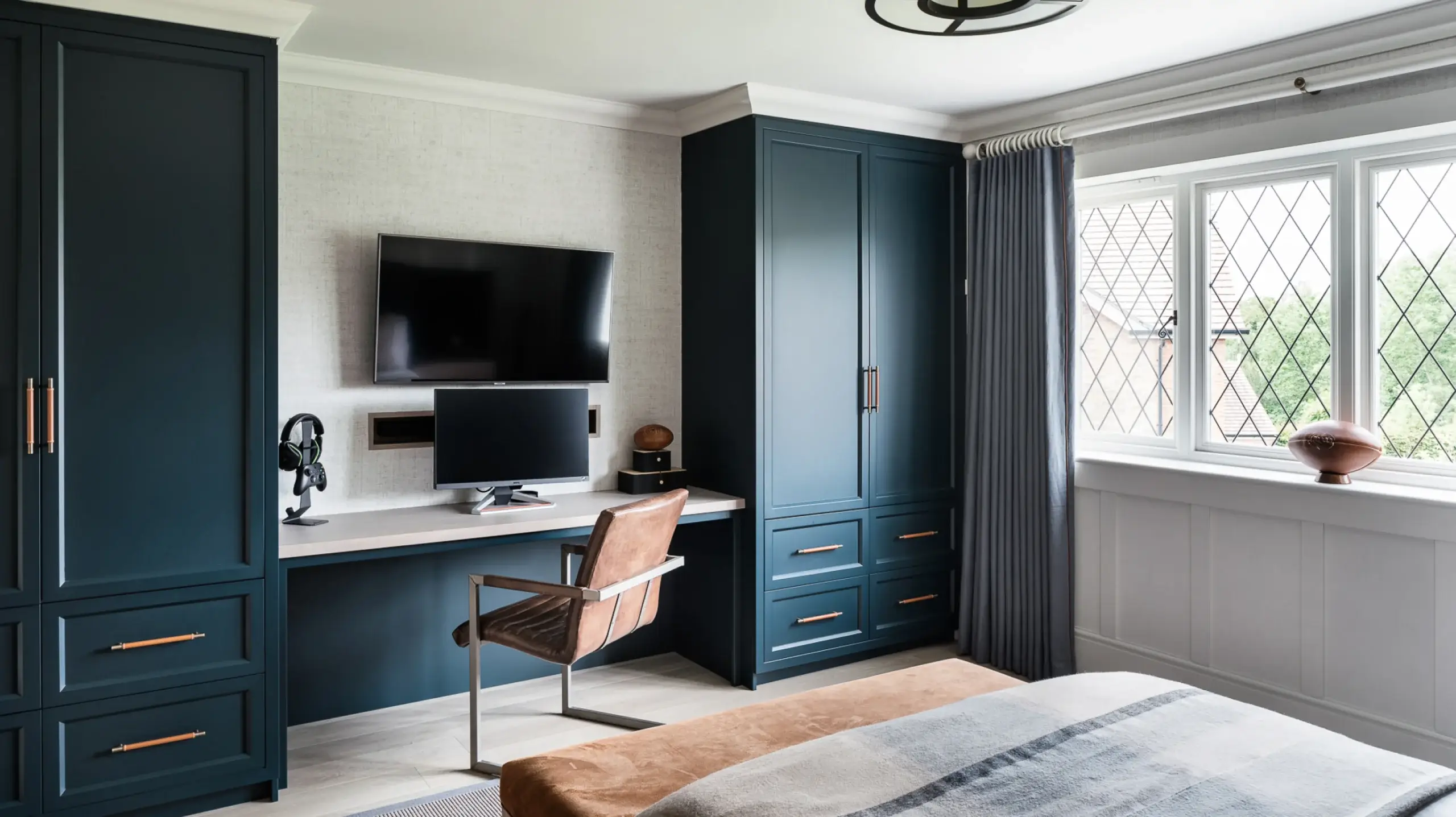 Bedroom workspace with dark teal built-in cabinets, desk, monitor and TV, leather chair, and diamond-pane window with curtains.