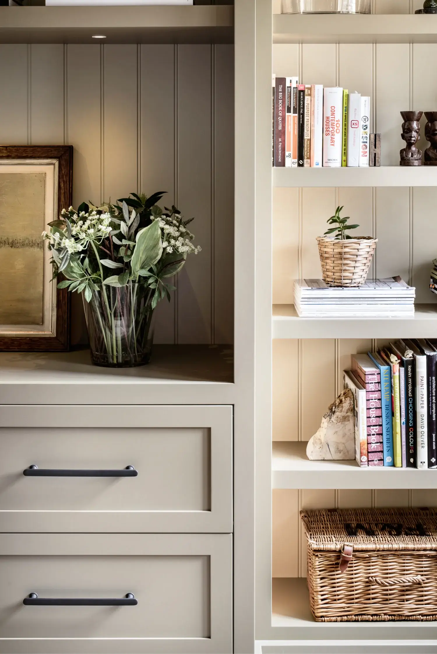 Built-in beige shelving with books, wicker basket, small potted plant, rock paper stack, and a vase of greenery beside framed art.