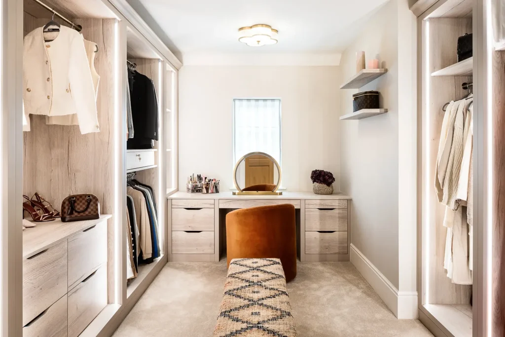 Walk-in dressing room with light wood built-in wardrobes, vanity with round mirror, rust velvet stool, and patterned bench.