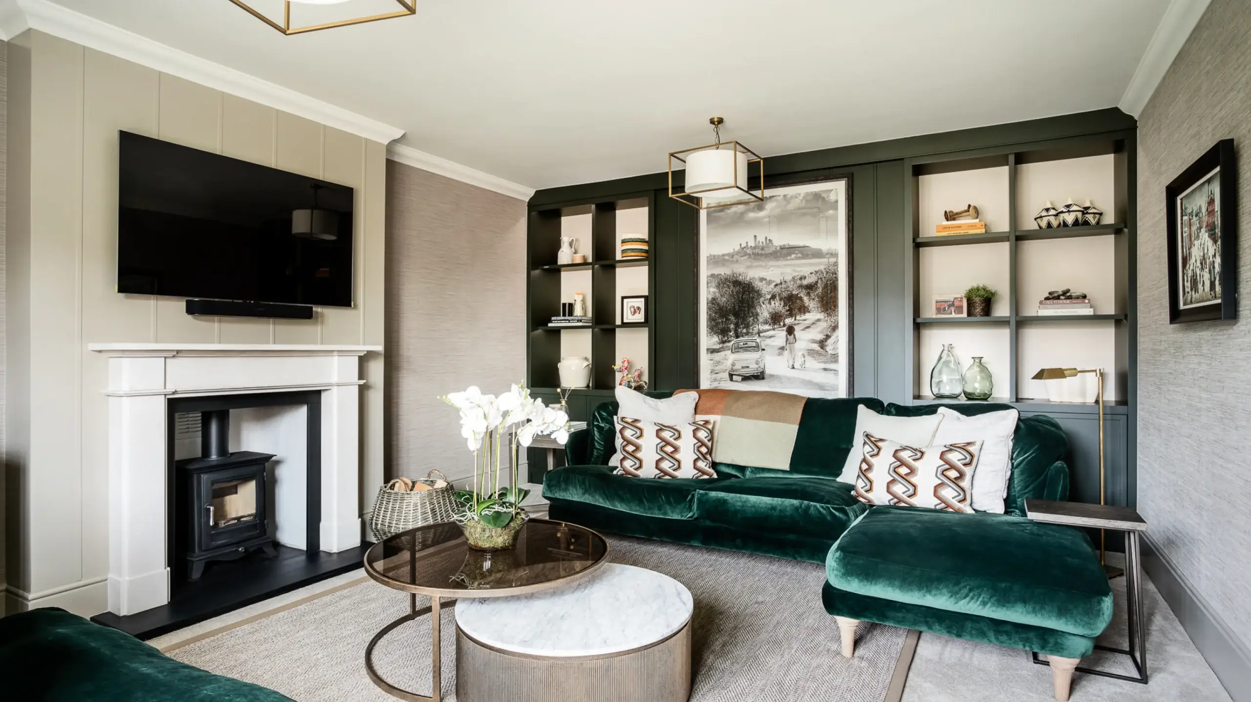 Living room with deep green velvet sectional and ottoman, marble-top and glass coffee tables, built-in dark shelves, and a white fireplace with wall-mounted TV.