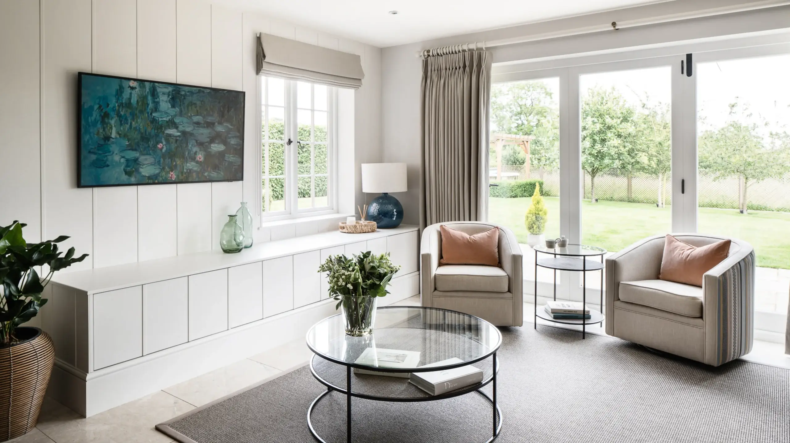 Bright living room with two beige armchairs, round glass coffee table, TV on paneled wall, and large garden-facing windows.