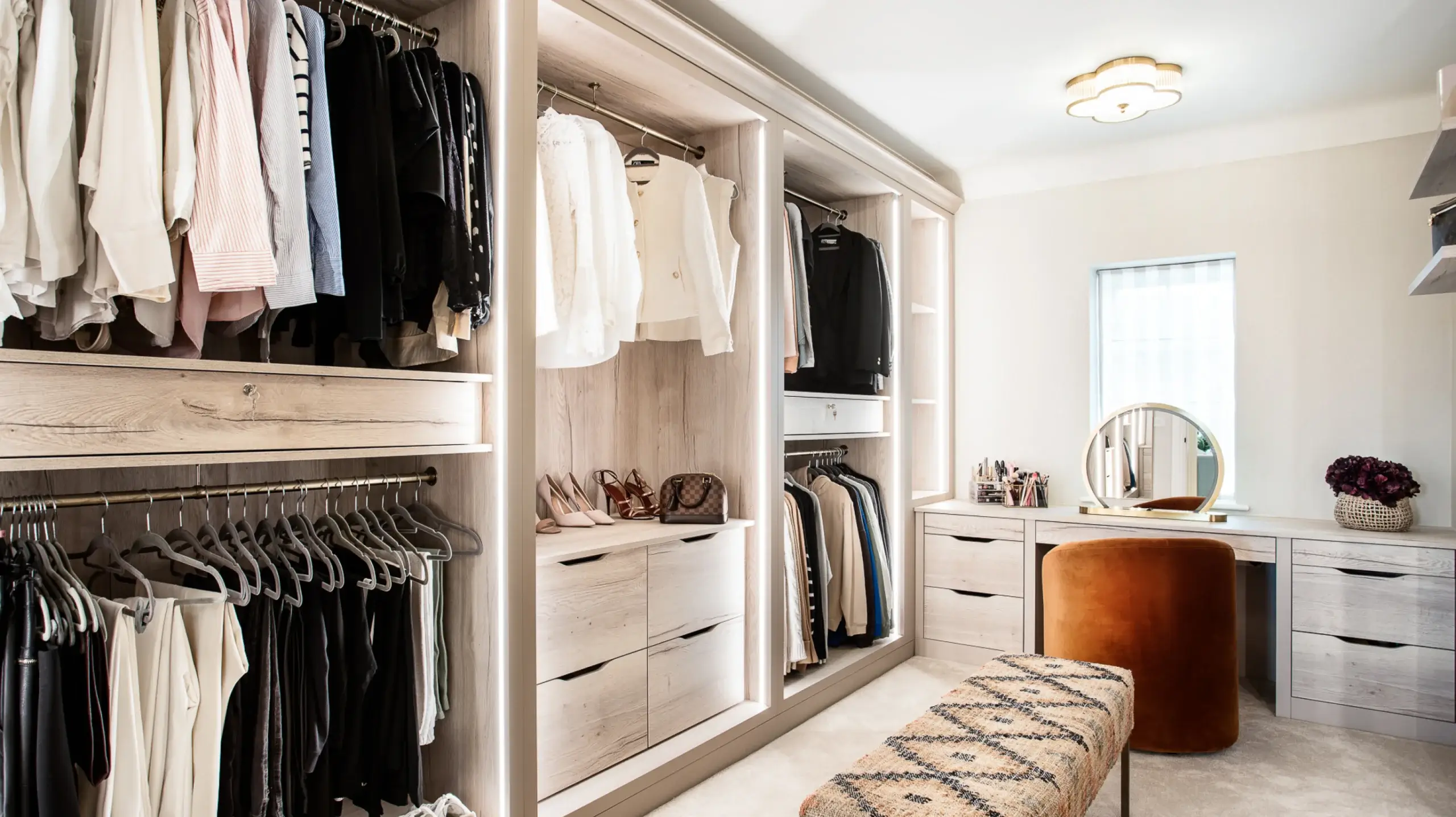 Organized walk-in closet with light wood shelving, hanging clothes, shoe storage, vanity with round mirror, and orange stool.