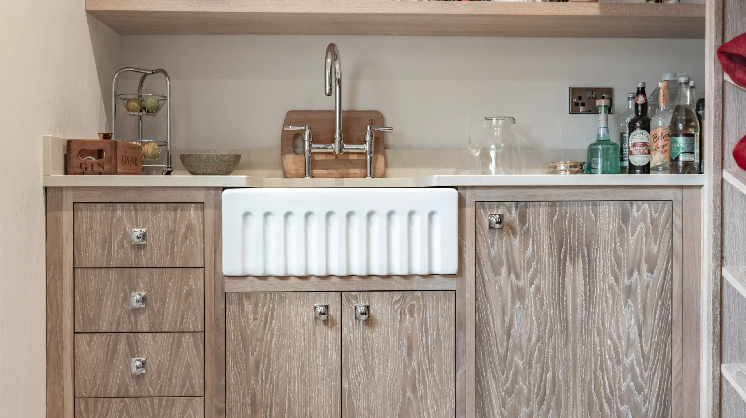 Kitchen sink with chrome faucet set into white farmhouse apron-front basin and light wood cabinets, counter with bottles and fruit rack