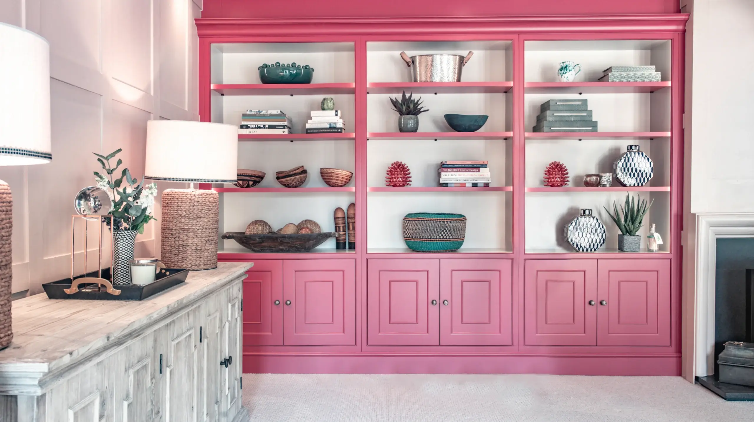 Built-in pink shelving unit filled with decorative bowls, books, baskets and plants beside a distressed white console with lamps.