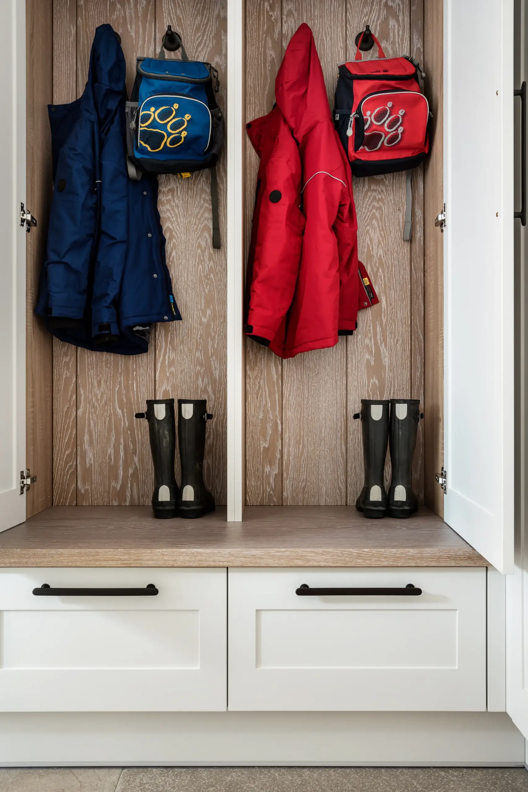Two open wood-backed cubbies with kids' raincoats (blue and red), matching backpacks hung above, and pairs of black rain boots below on a bench with white drawers.