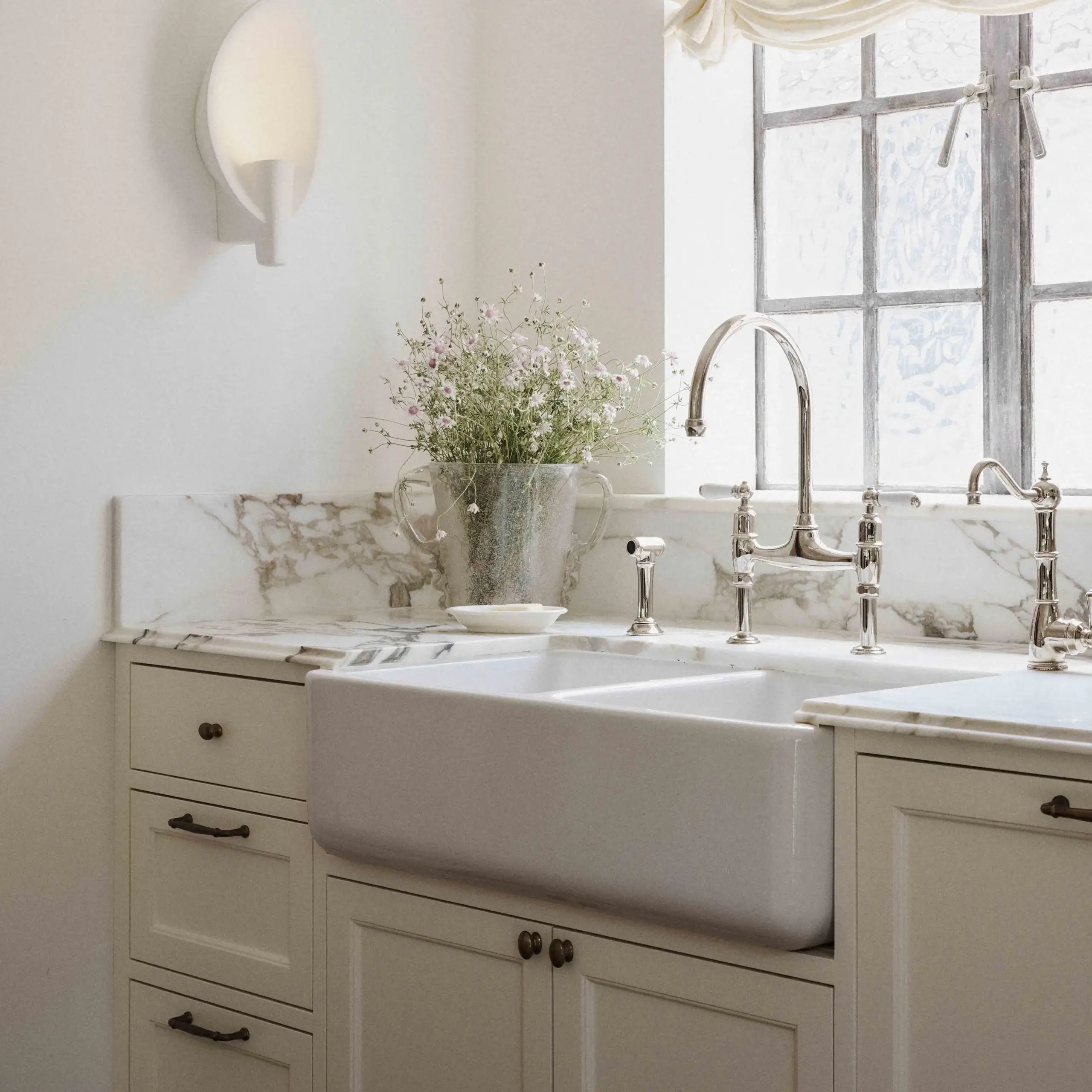 Farmhouse-style white ceramic apron sink with chrome faucet, marble countertop, and a vase of wildflowers by a window