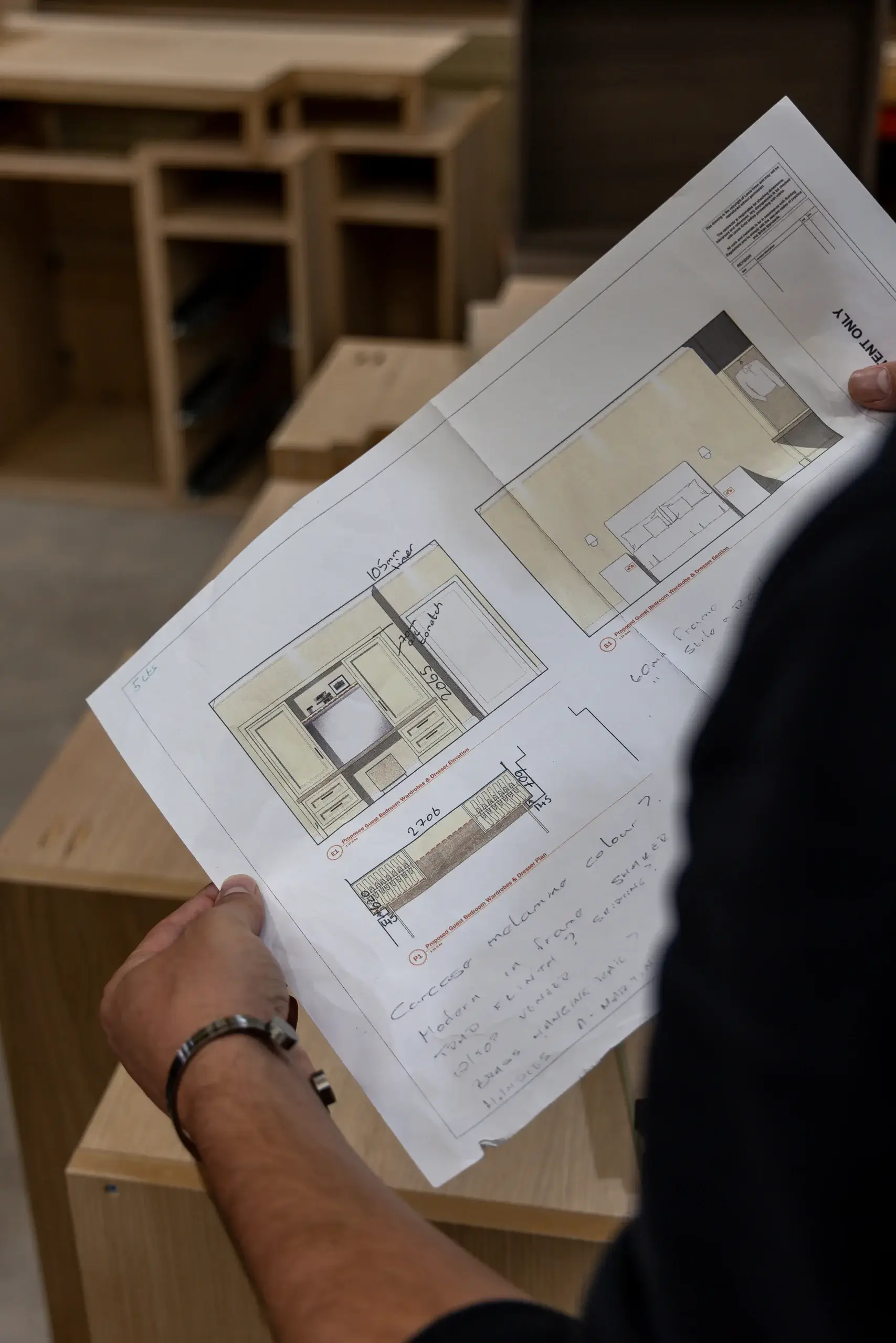 Hands holding a printed furniture plan with cabinet elevations and handwritten notes, set over unfinished wooden cabinet pieces.