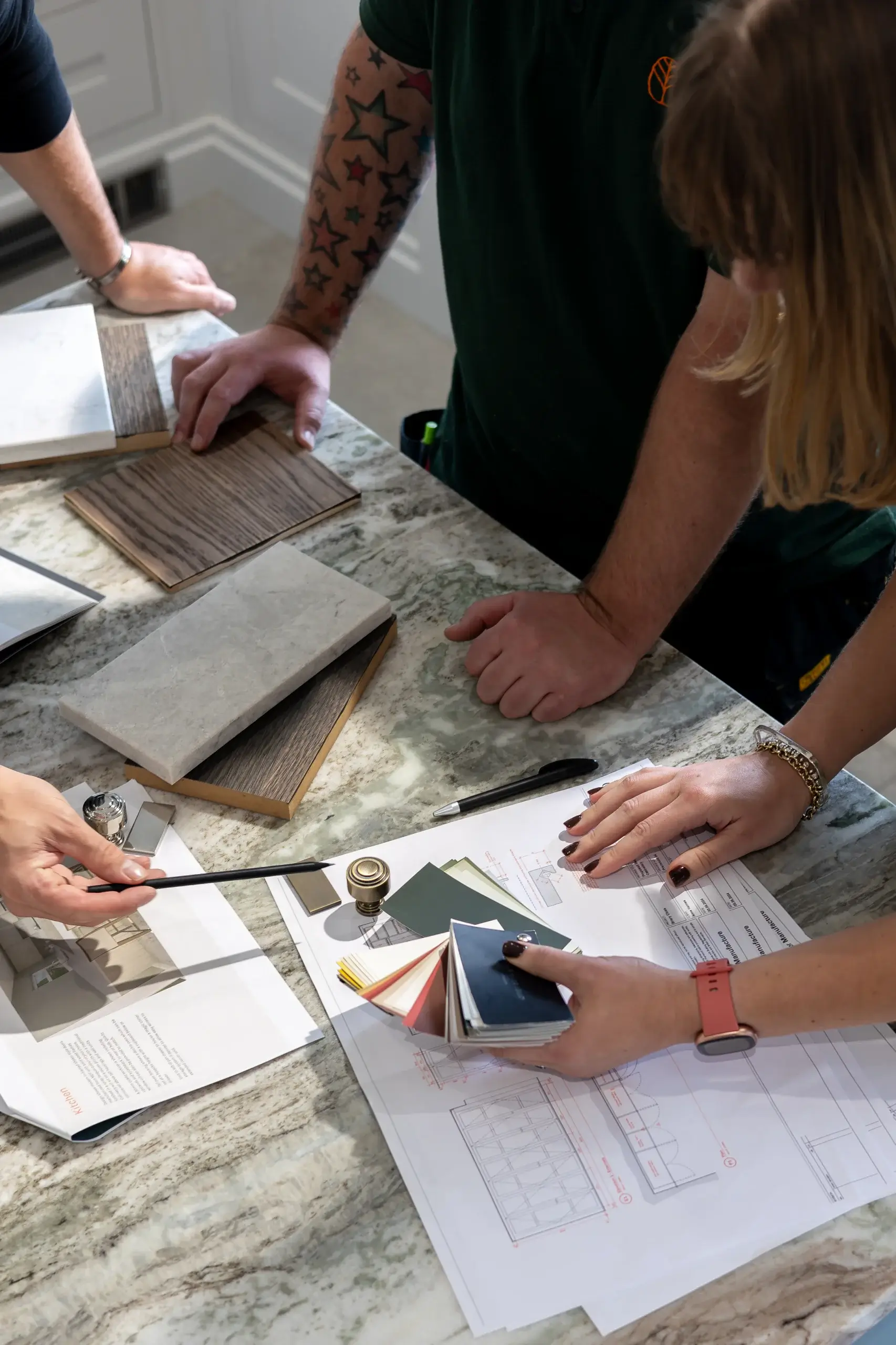 Hands of three people examining paint swatches, wood and tile samples and architectural plans on a marble countertop.