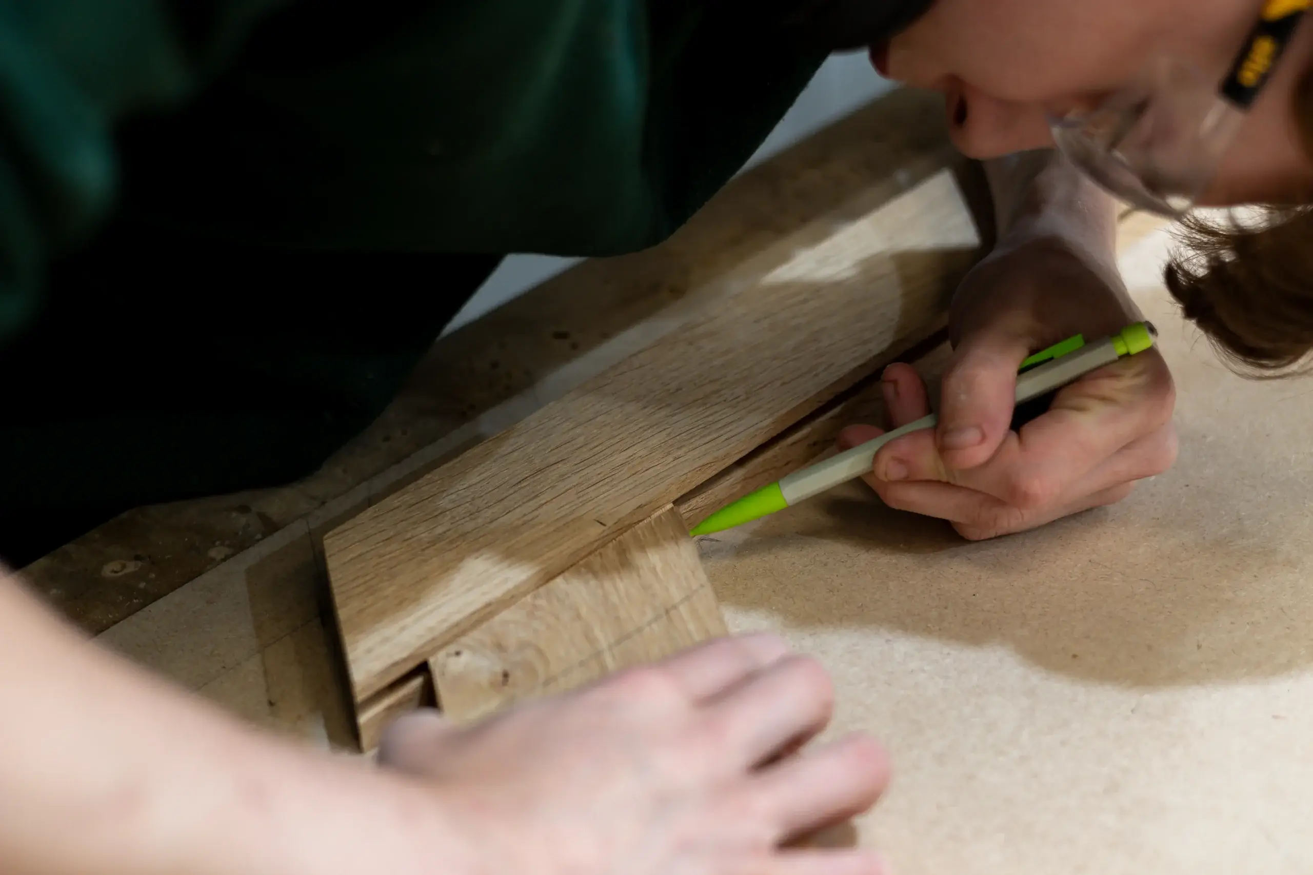 Person marking a groove on a wooden board with a green-capped pencil while wearing safety glasses and leaning close to the workpiece.