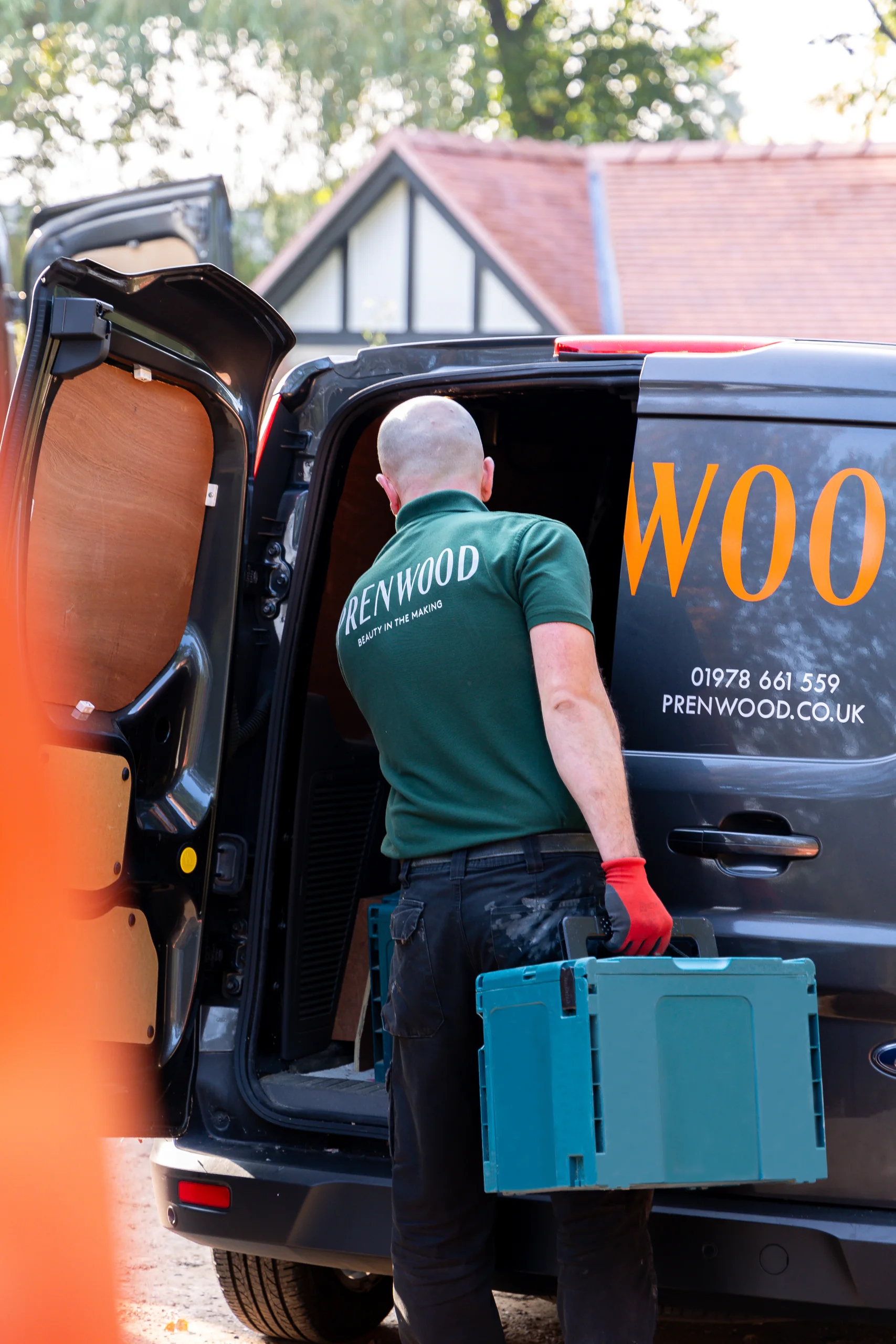 Worker in green "Prenwood" polo loading a blue toolbox into a company van with orange lettering.