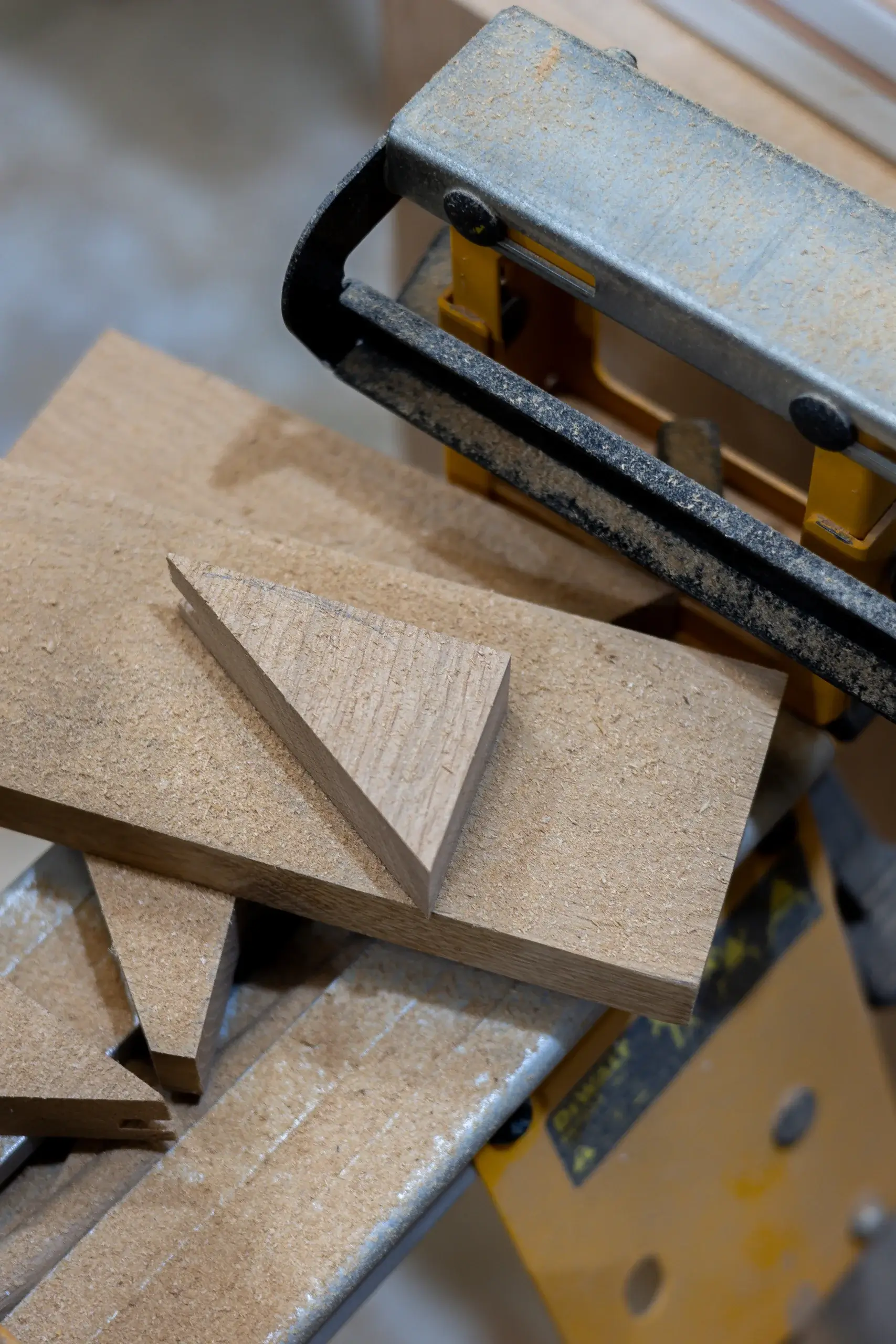 Small triangular wood wedge and rectangular wood pieces covered in sawdust next to a yellow and metal woodworking vise.