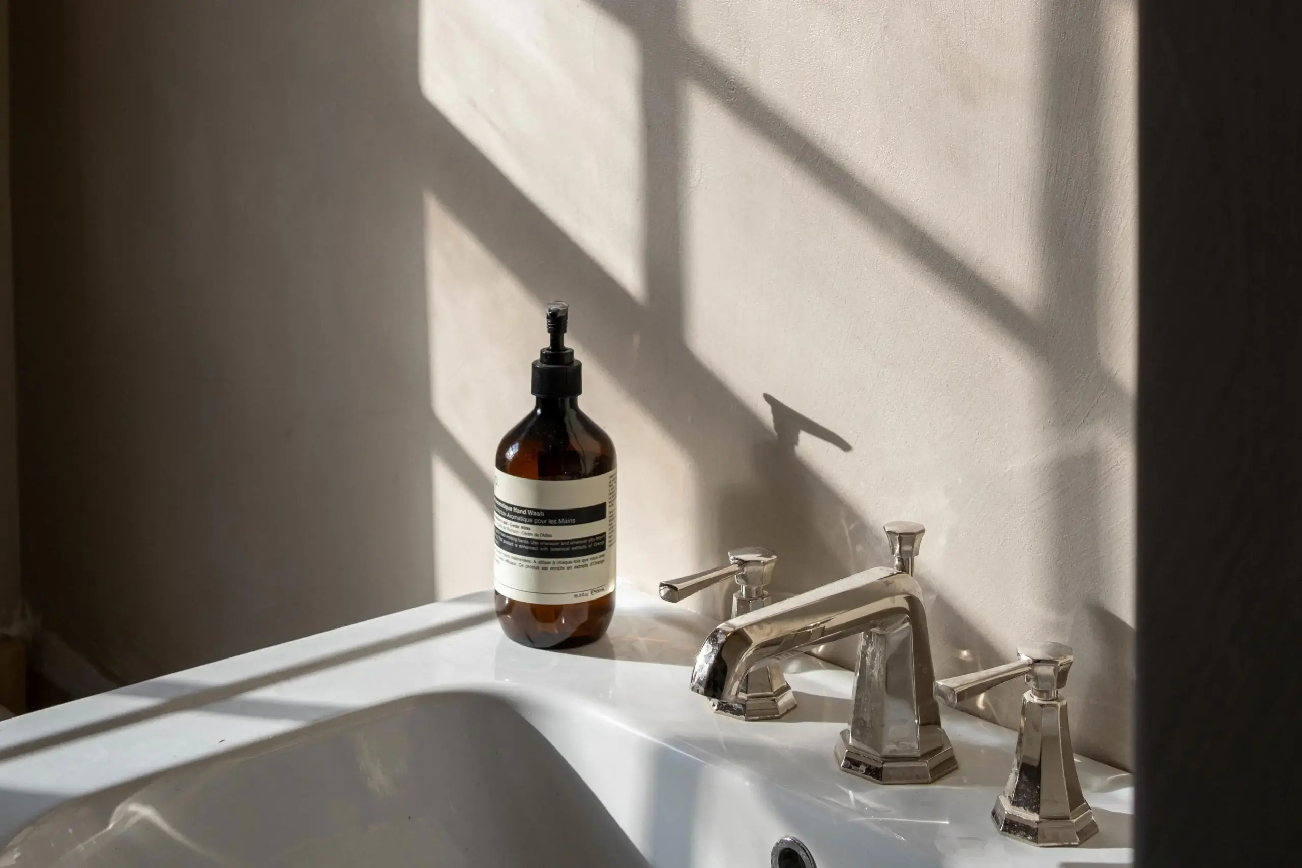 Chrome bathroom faucet and handles on a white sink next to an amber pump soap bottle casting window grid shadows on a beige wall
