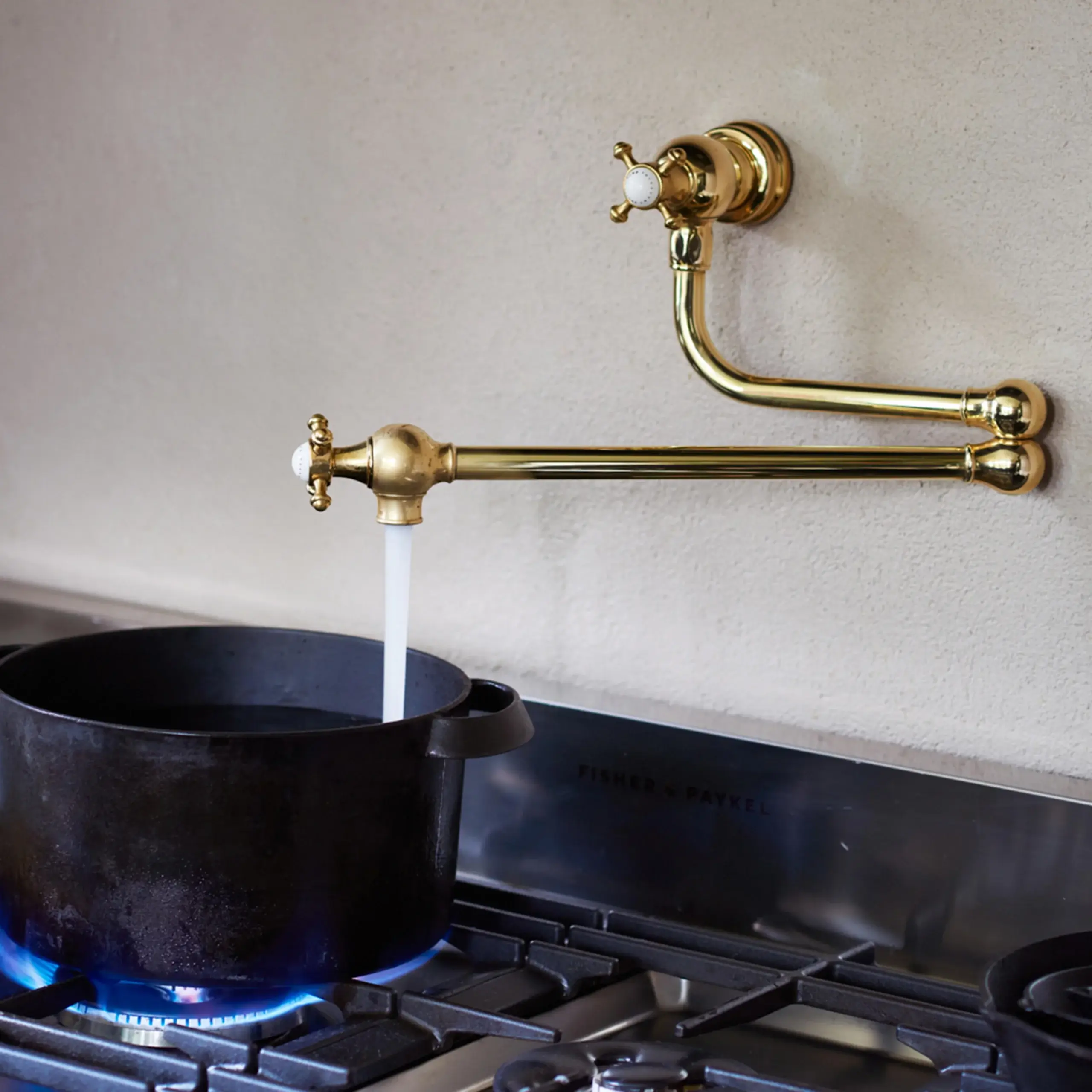 Brass pot filler faucet mounted above a gas stove pouring water into a black cast-iron pot with blue flame beneath.