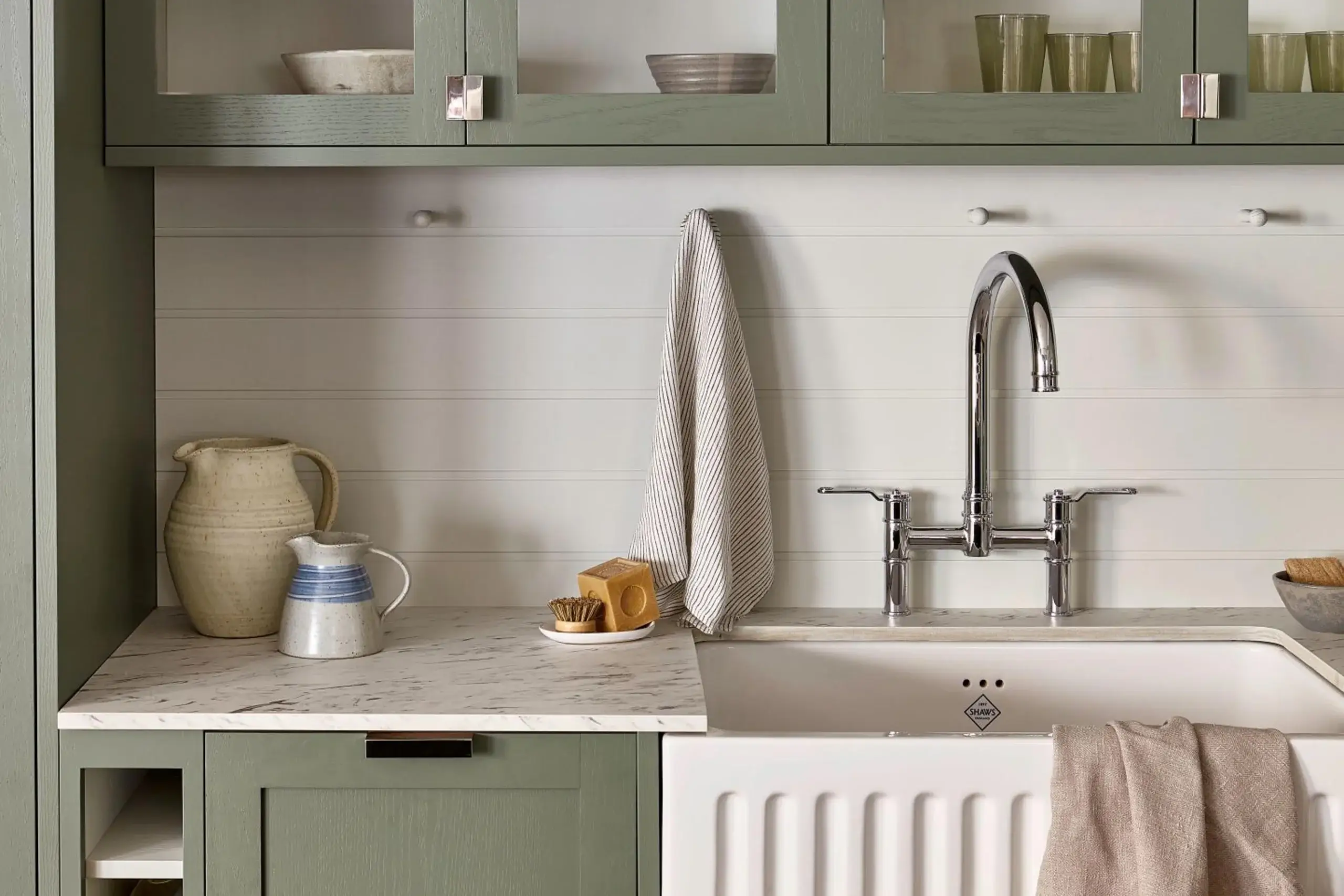 Farmhouse kitchen sink area with chrome gooseneck faucet, white apron-front sink, green cabinets, striped dish towel, and ceramic pitchers on marble countertop