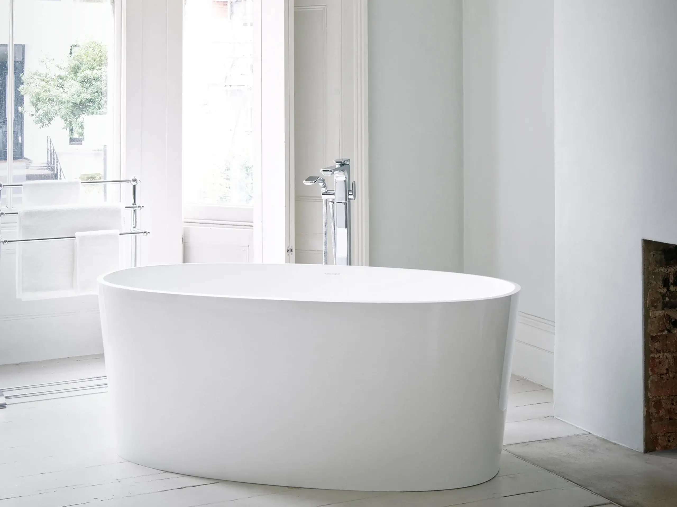 Freestanding white oval bathtub in a bright minimalist bathroom with chrome floor-mounted faucet and towel rack