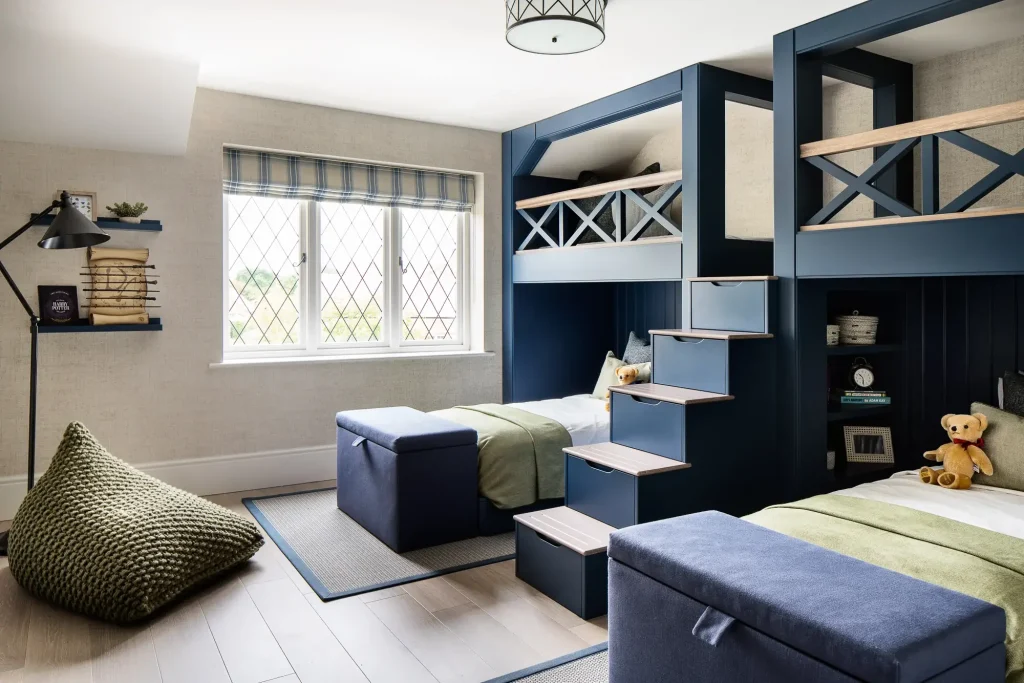 Children's bedroom with navy bunk beds, built-in stair drawers, green knit beanbag, and twin beds with teddy bears.