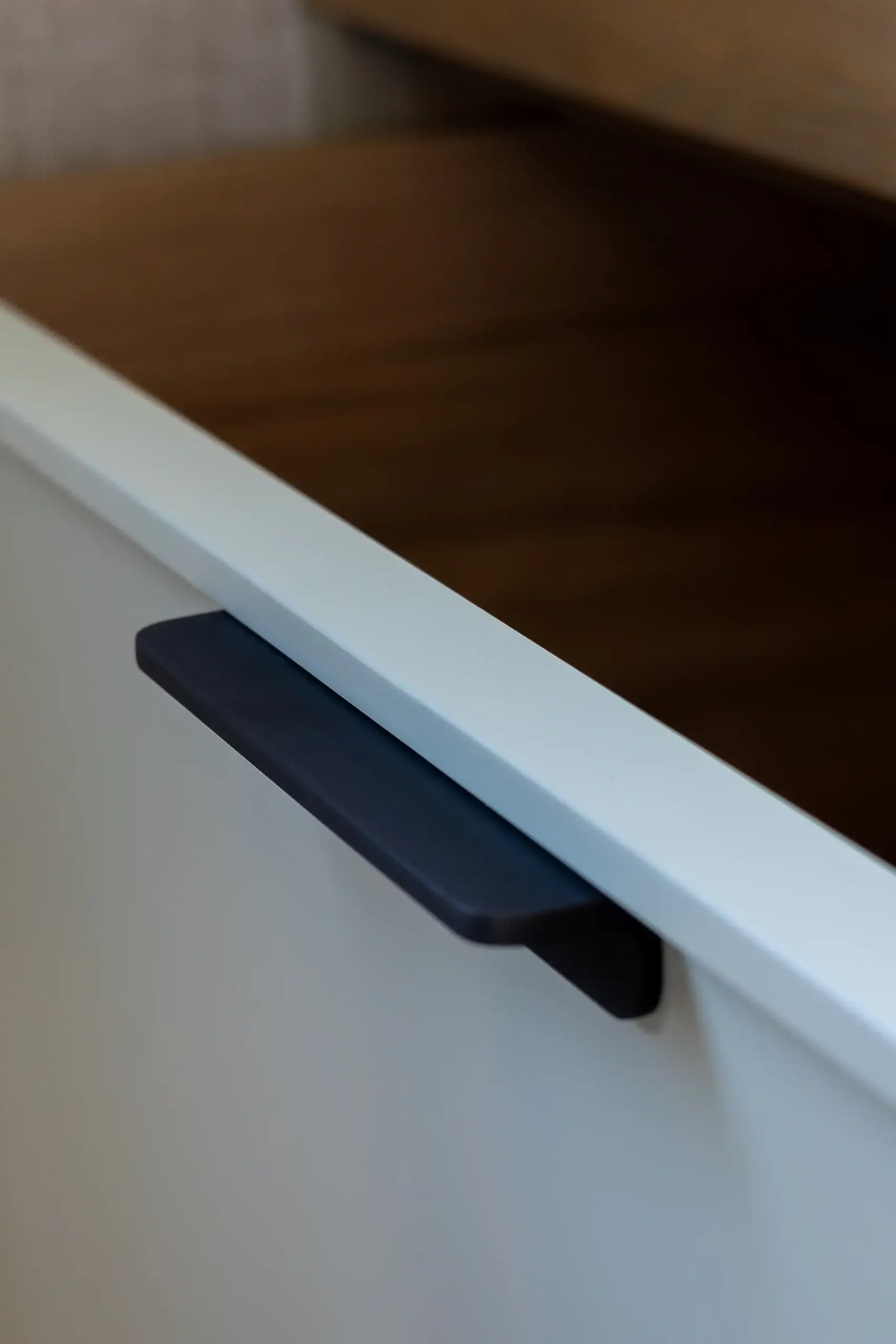 Slim matte black drawer pull mounted on a white cabinet front seen at an angle, with a blurred wood surface in the background.