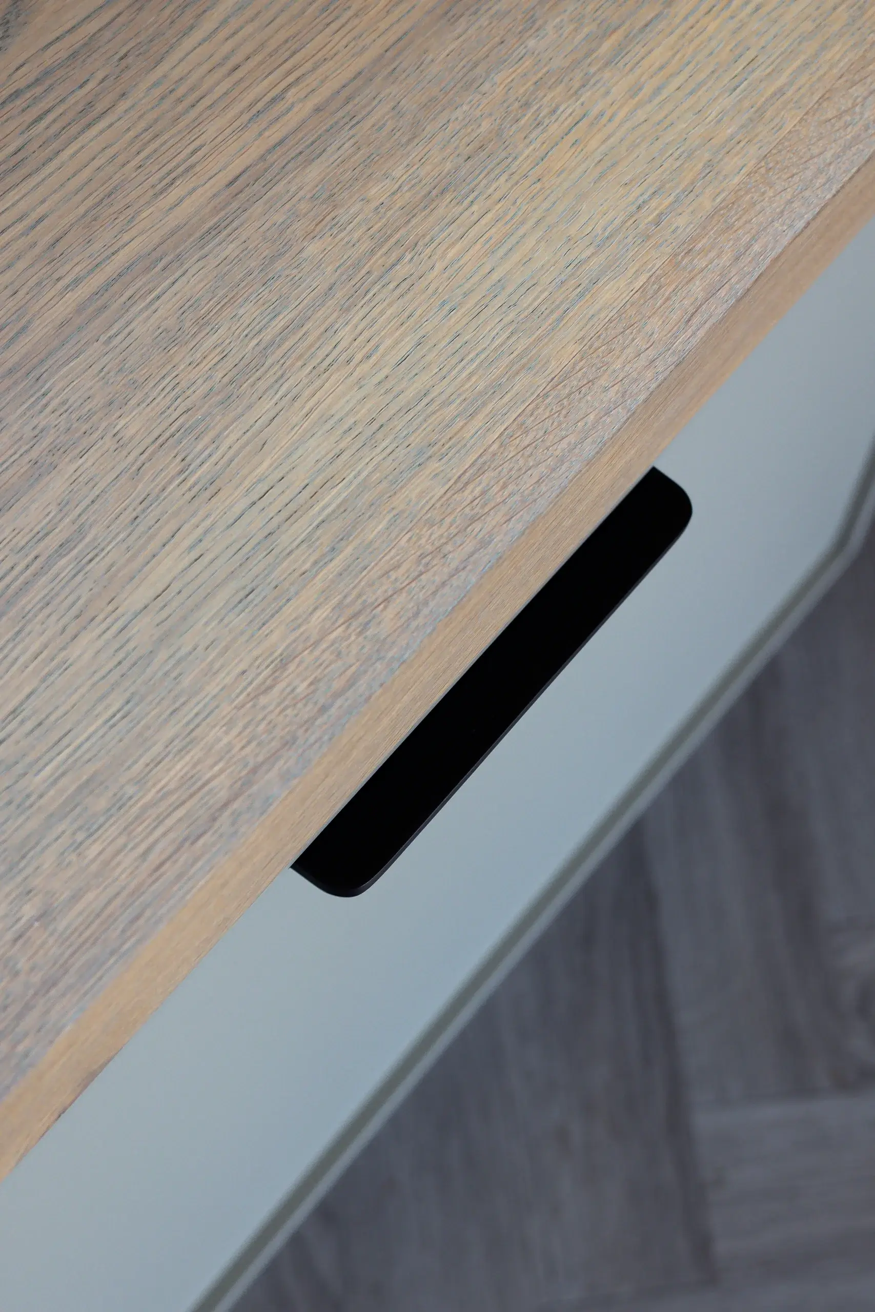 Close-up of a light wood tabletop edge with a black recessed drawer handle and gray floor visible below.