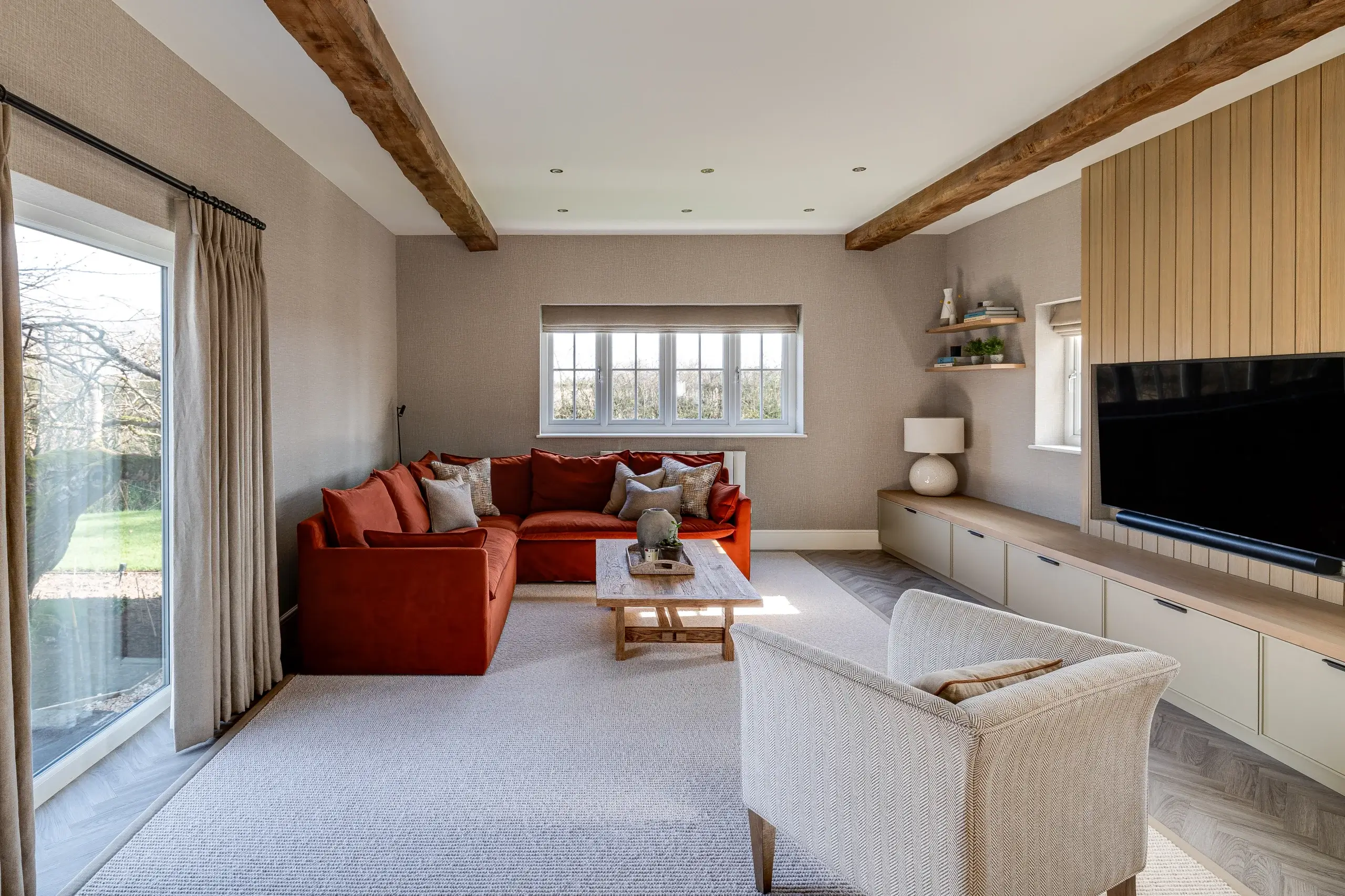 Bright living room with an orange sectional, wooden coffee table, beige armchair, built-in media unit and exposed ceiling beams