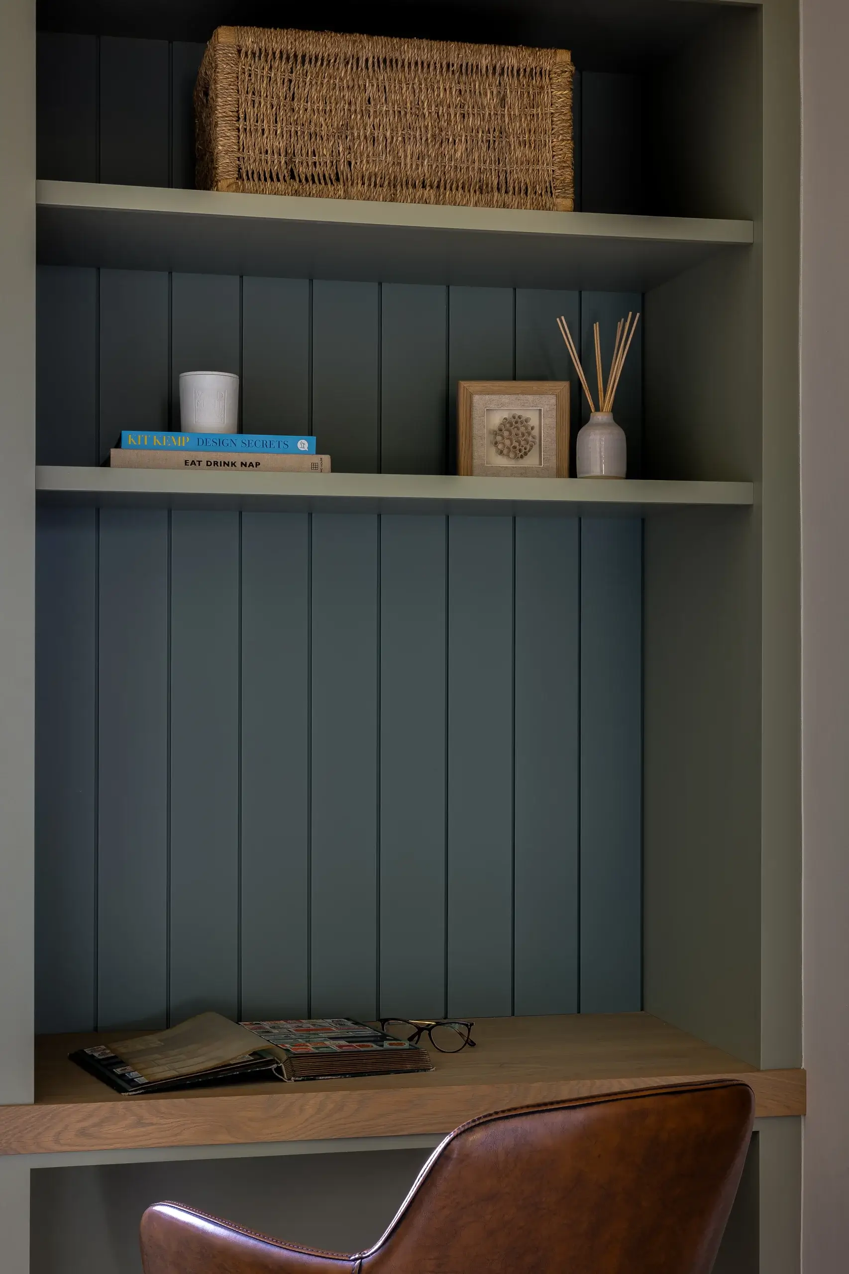 Built-in desk nook with teal vertical shiplap, wooden shelf holding wicker basket, books, framed art, reed diffuser, and leather chair.