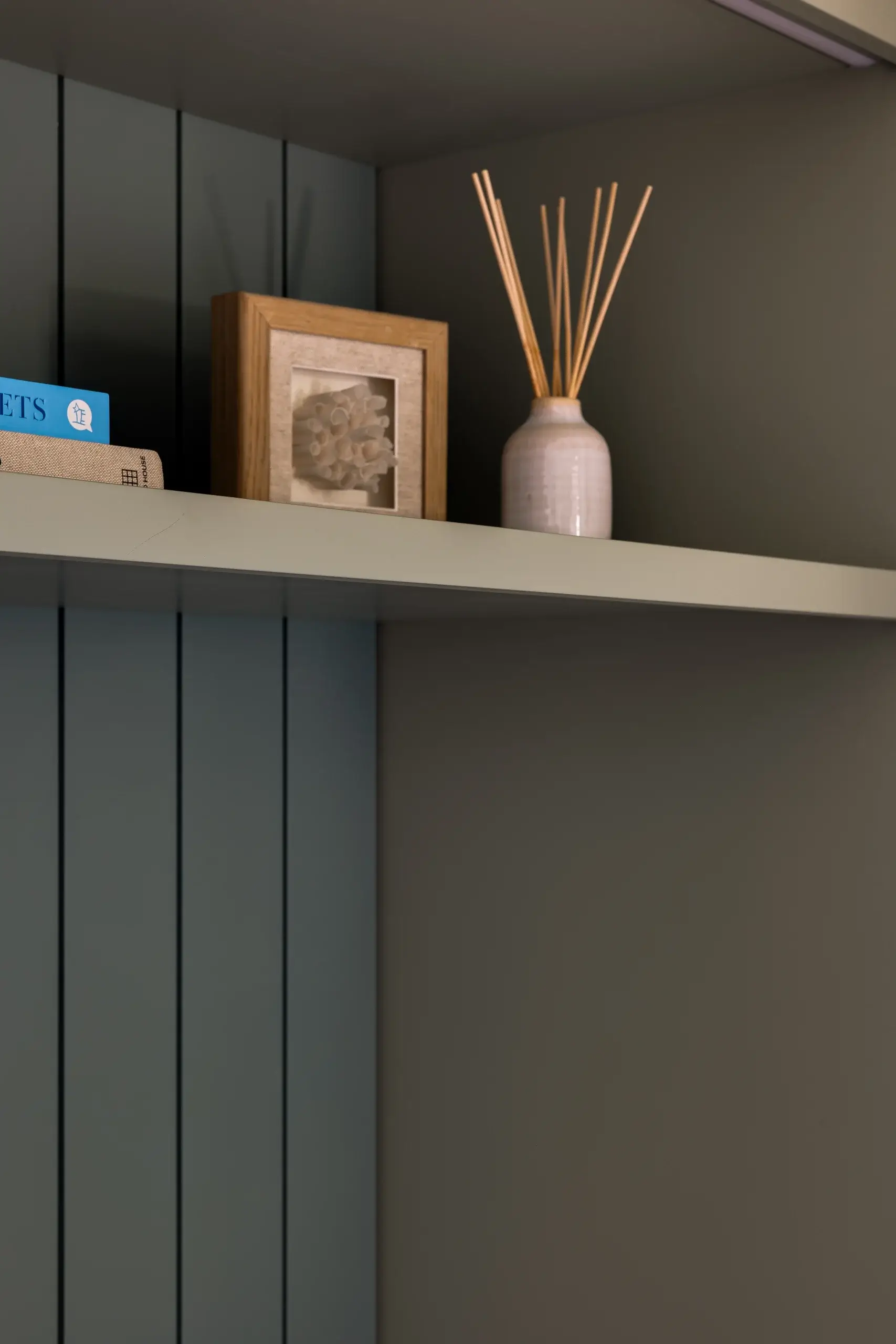 Wooden shelf holding a small framed coral sculpture and a ceramic diffuser vase with tall reed sticks against paneled wall.
