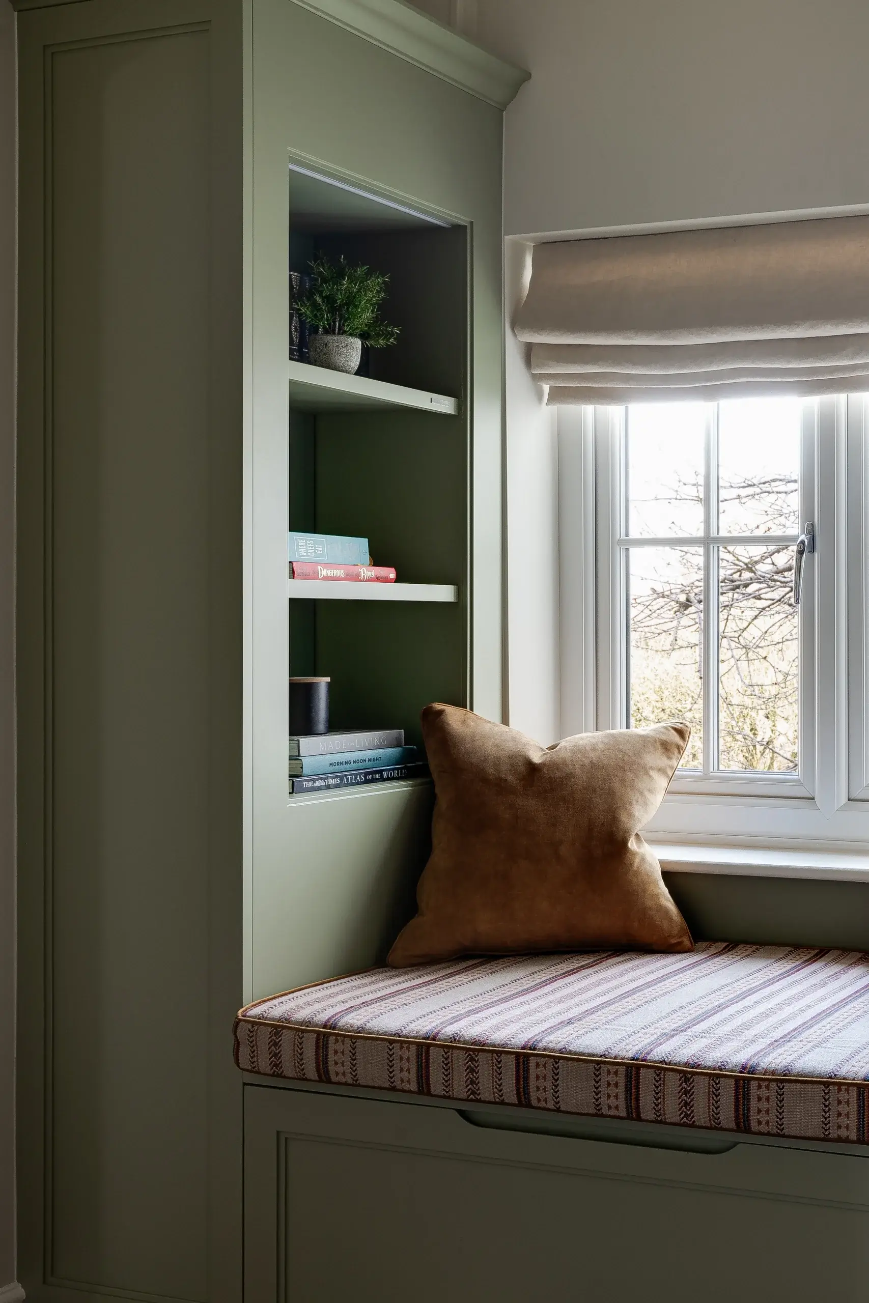 Cozy window seat with striped cushion, brown velvet pillow, built-in green shelving holding books and a small potted plant.
