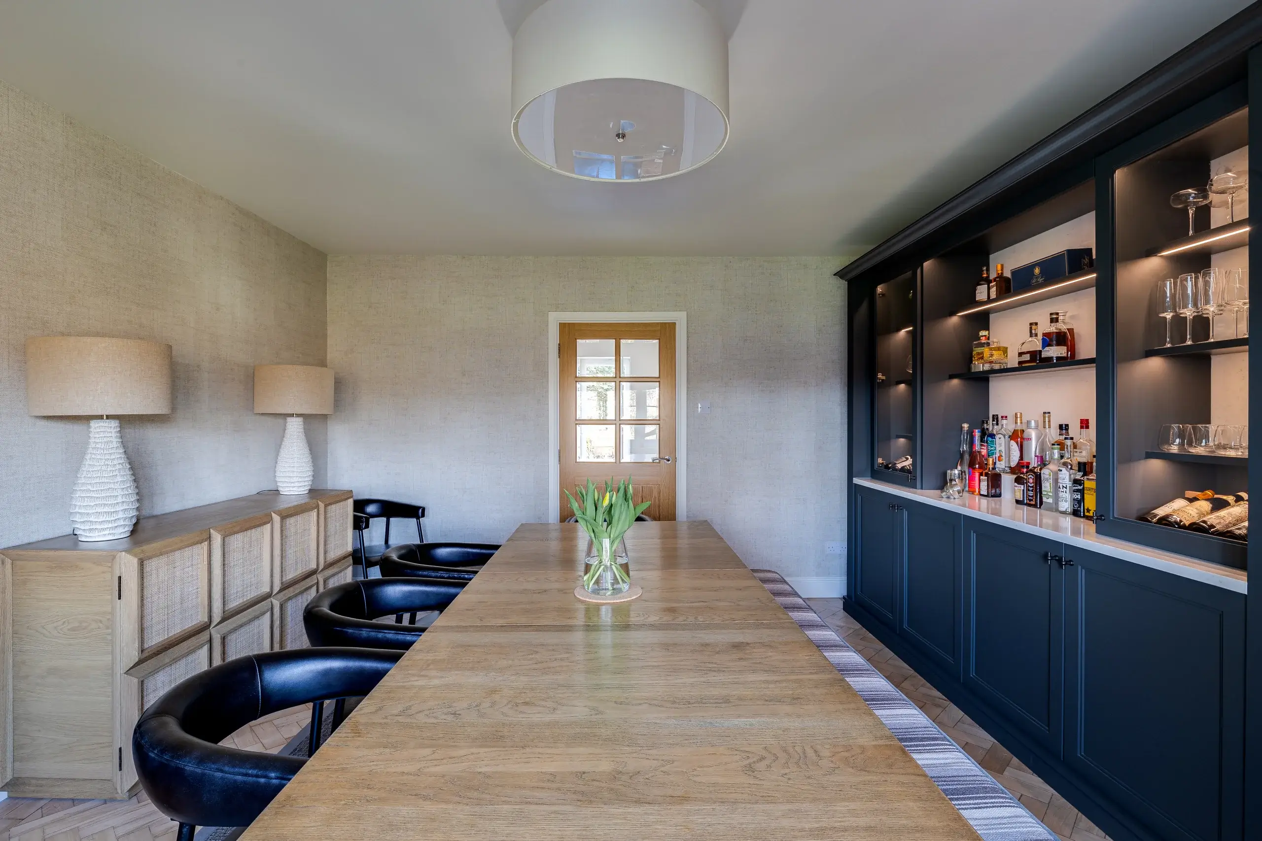 Long wooden dining table with tulip vase, black leather chairs, sideboard with lamps on left and dark built-in bar cabinets on right.