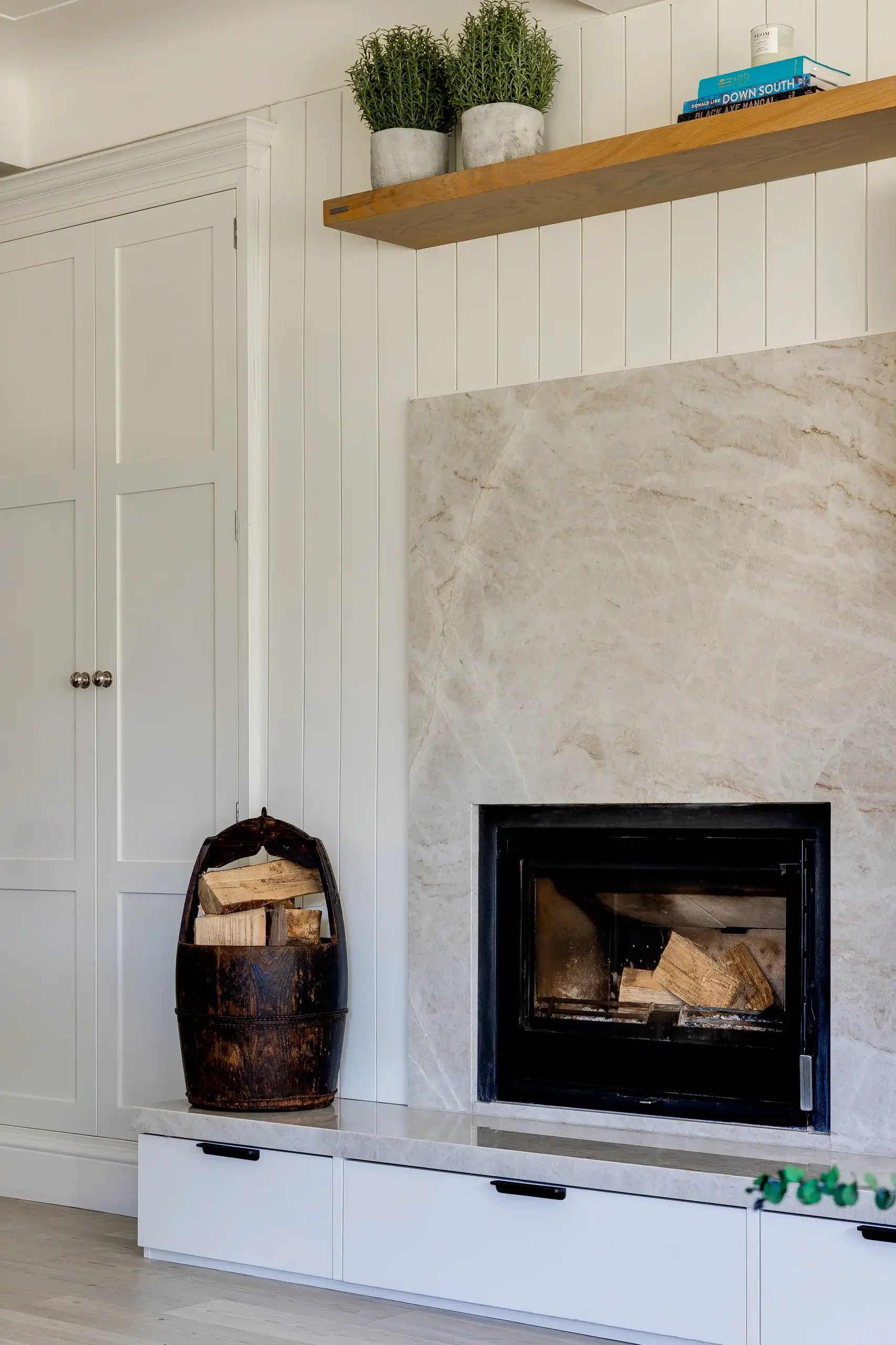 Built-in modern fireplace with marble surround, stacked firewood in a rustic bucket, white shiplap walls and a wooden shelf with plants.