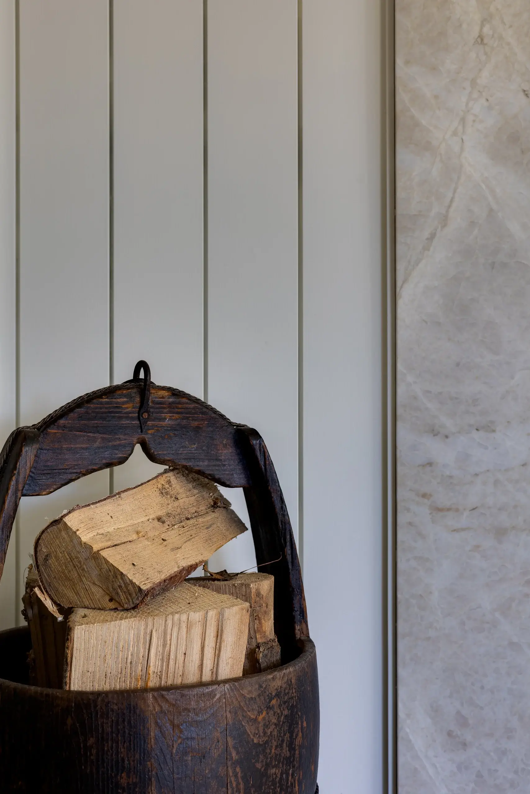 Stacked split firewood inside a dark wooden bucket with a curved handle against painted vertical panels.