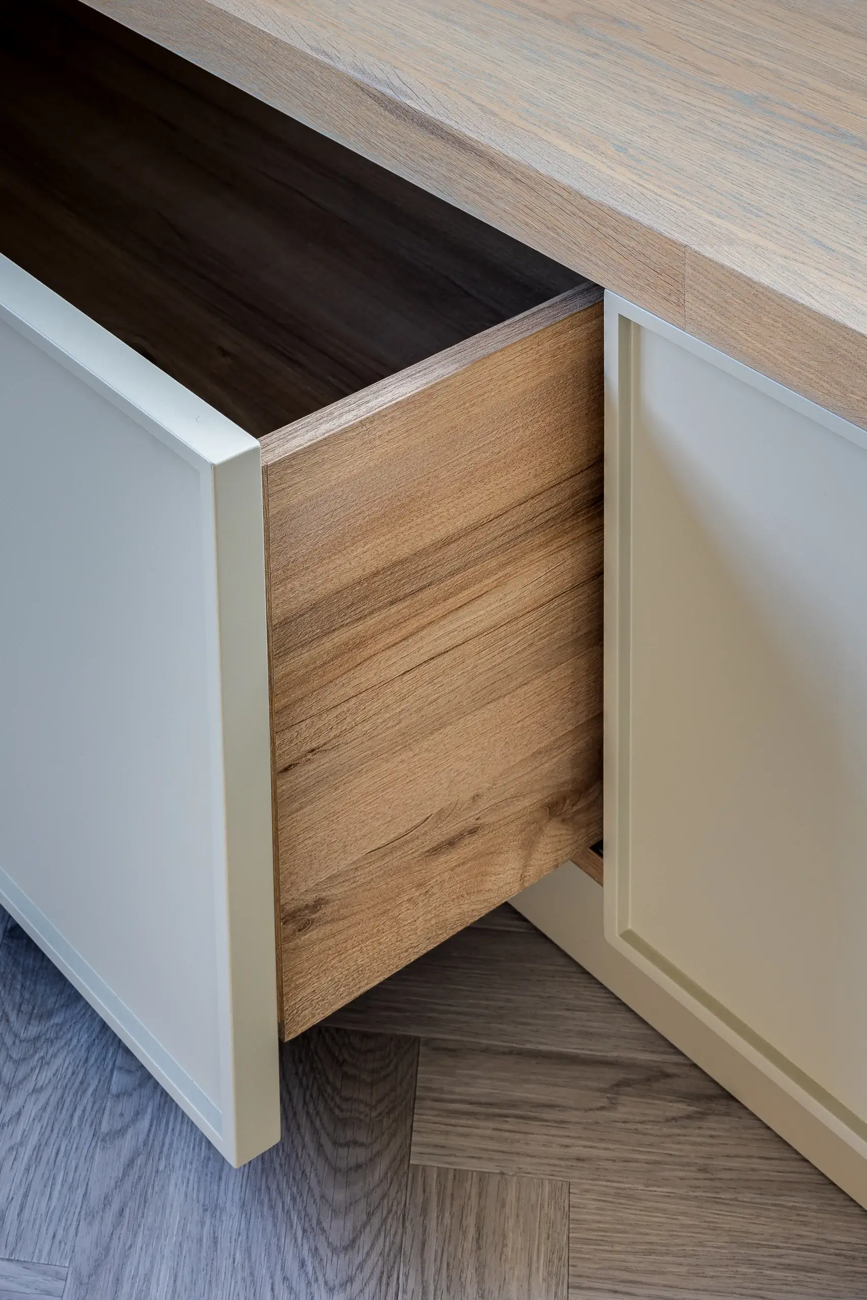 Partially open light wood drawer with white cabinet doors on a parquet wood floor, showing wood grain and corner details.